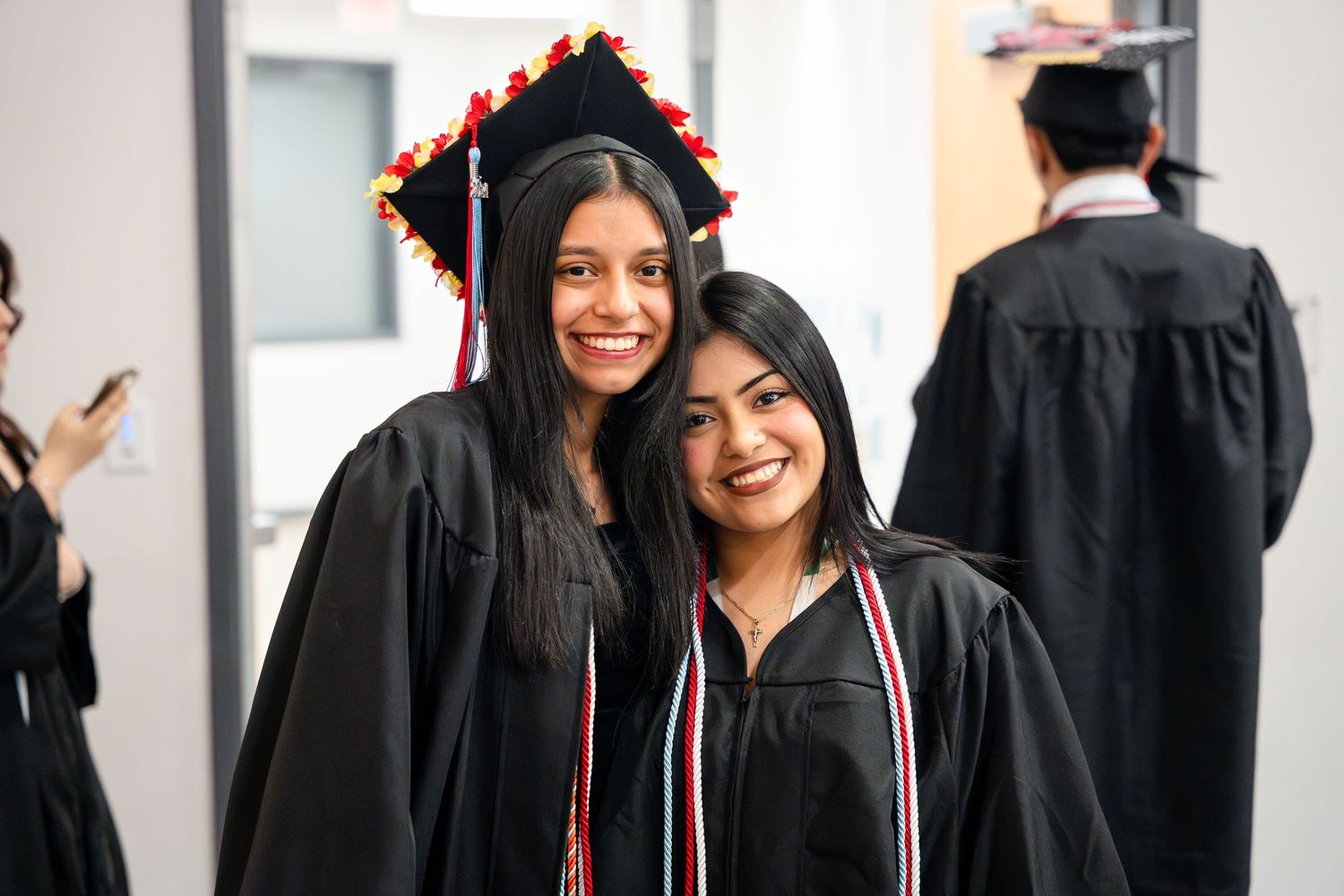 Two graduates in black caps and gowns smiling together during graduation ceremony.