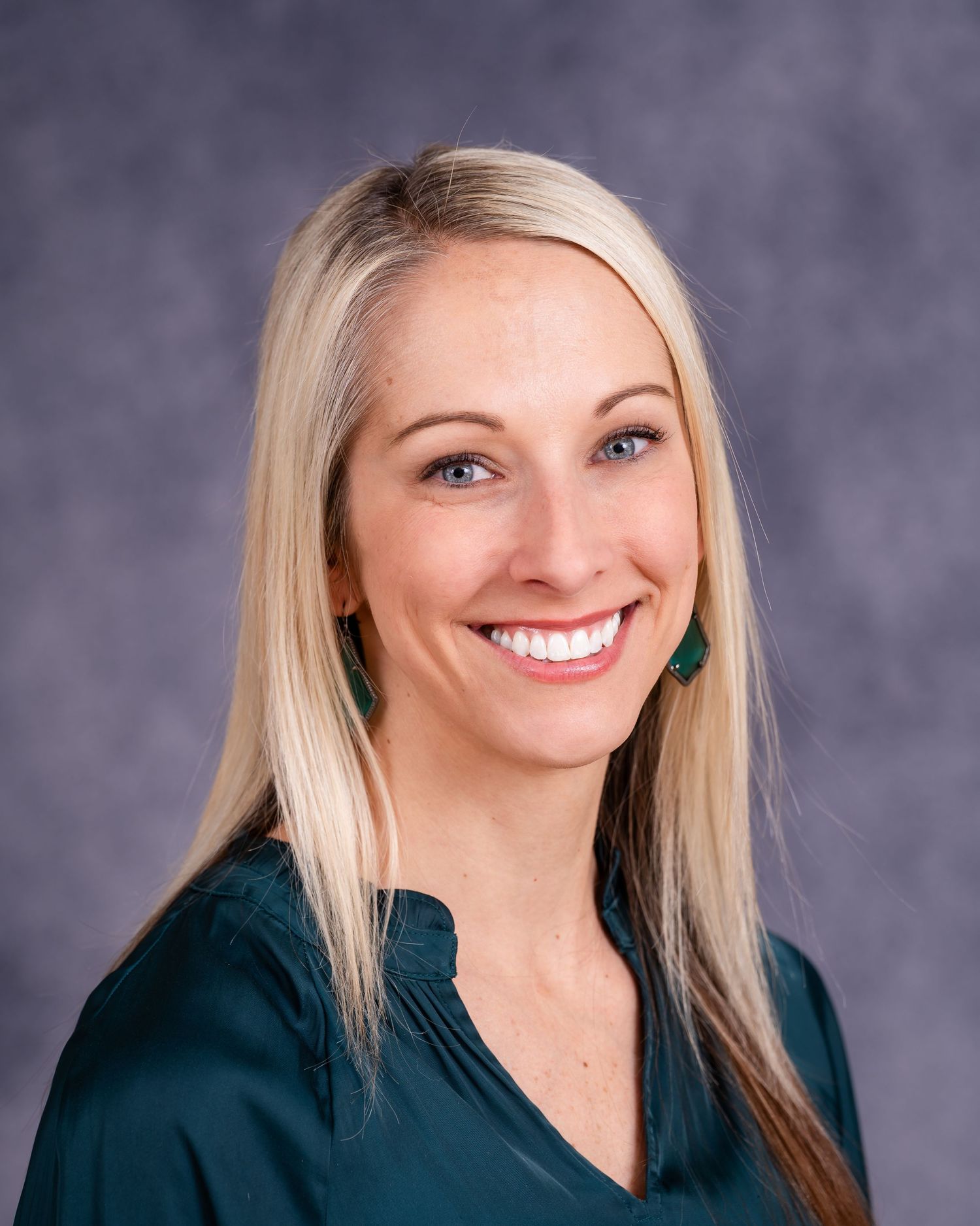 Professional headshot of woman in dark green blazer smiling against gray backdrop.