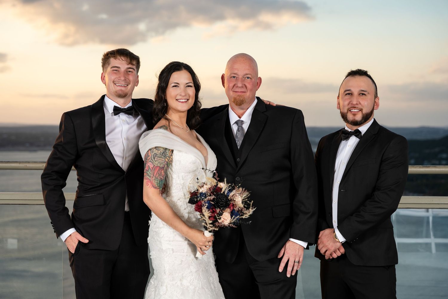 Formal group photo of wedding party members against sunset sky.