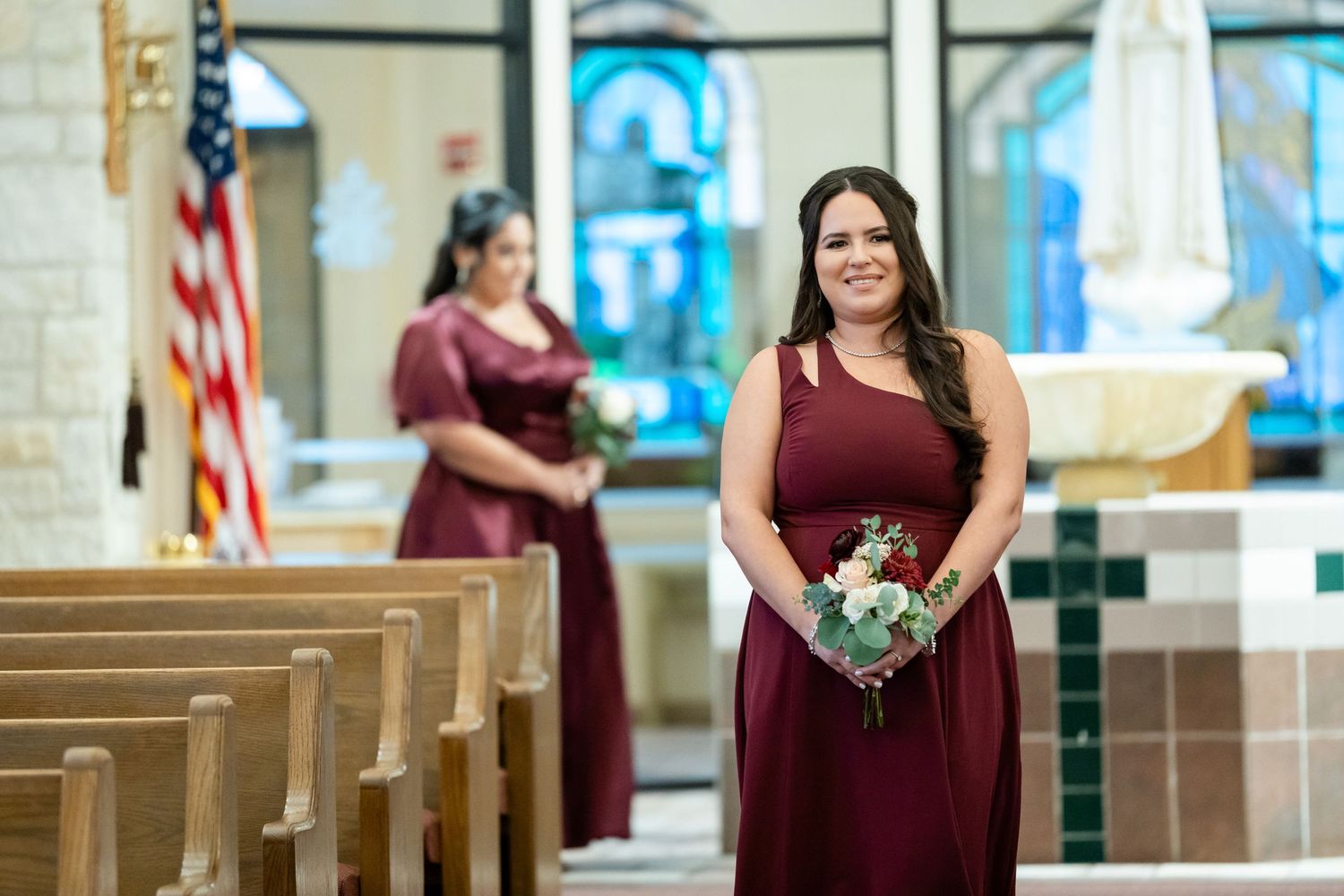 Bridesmaid in burgundy dress holds bouquet while standing in church aisle.