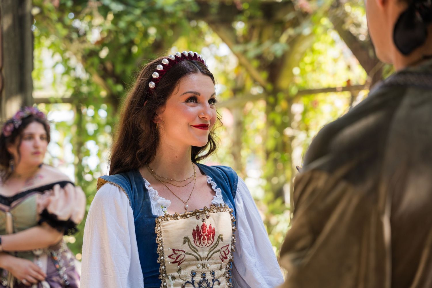 Woman in medieval dress with white blouse and blue bodice smiles during renaissance fair performance.