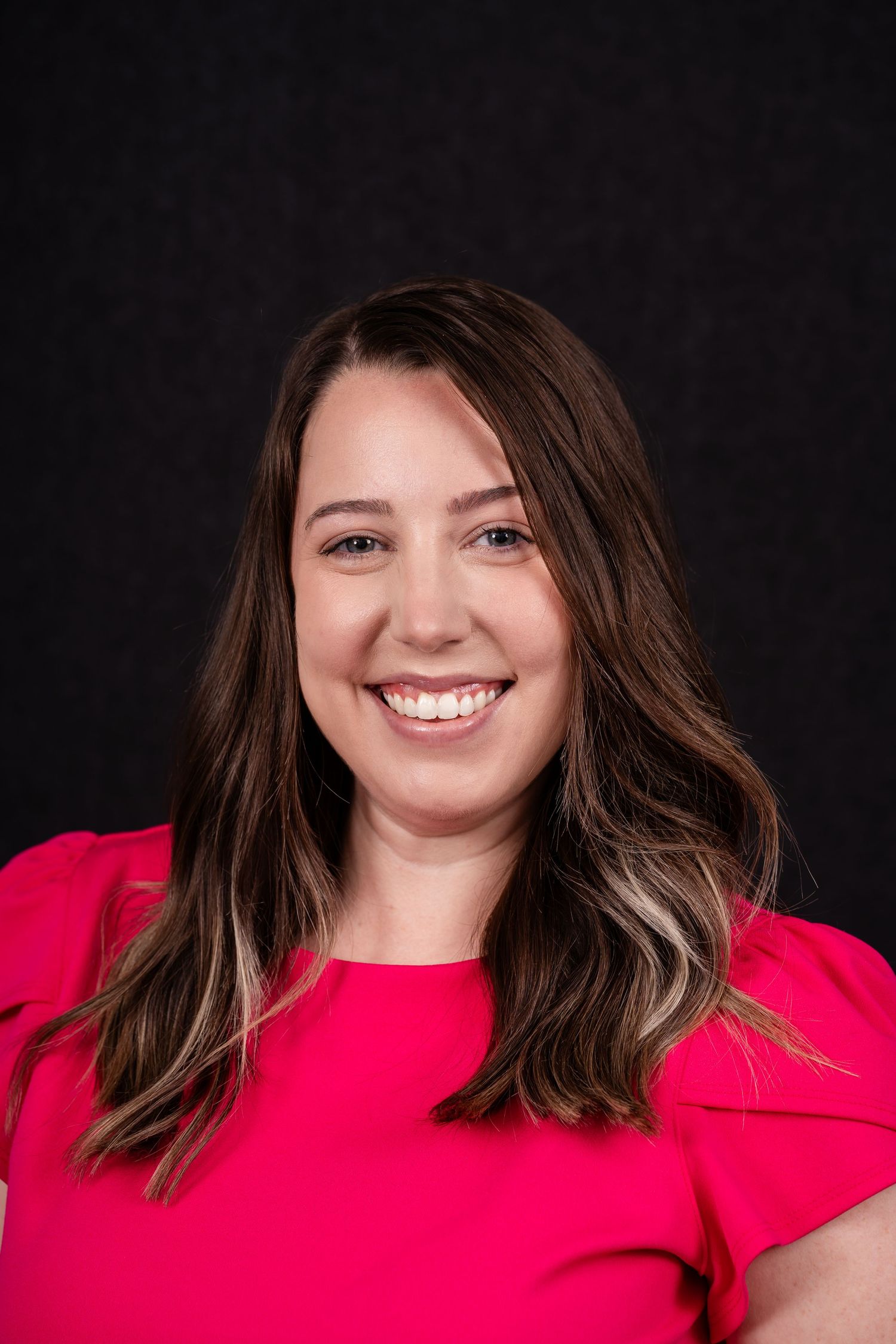 Professional headshot of someone in a bright red top against a dark background, with long brown hair and a warm smile.