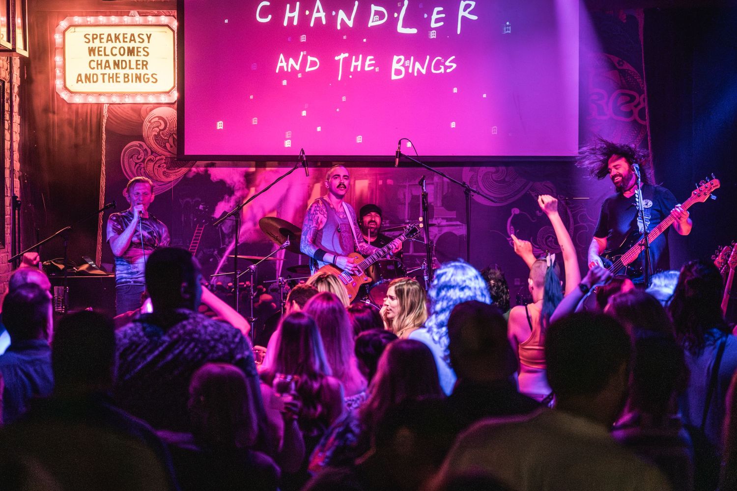 A band performs under pink lighting with an excited crowd at a venue with exposed brick walls.