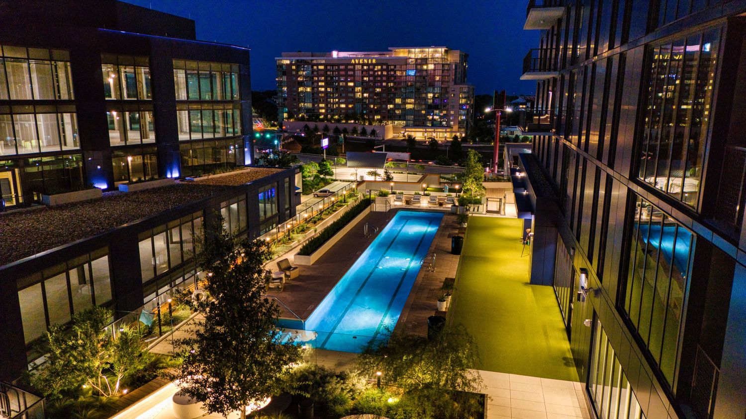 Evening photograph of blue-lit rooftop swimming pool flanked by modern residential buildings.