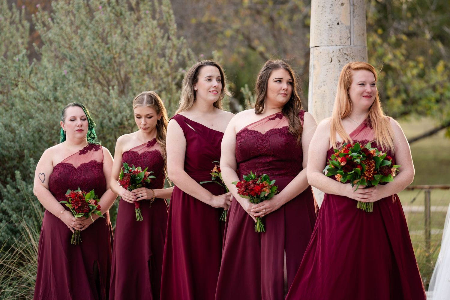 Bridesmaids in burgundy dresses hold bouquets while standing in a row at an outdoor venue.