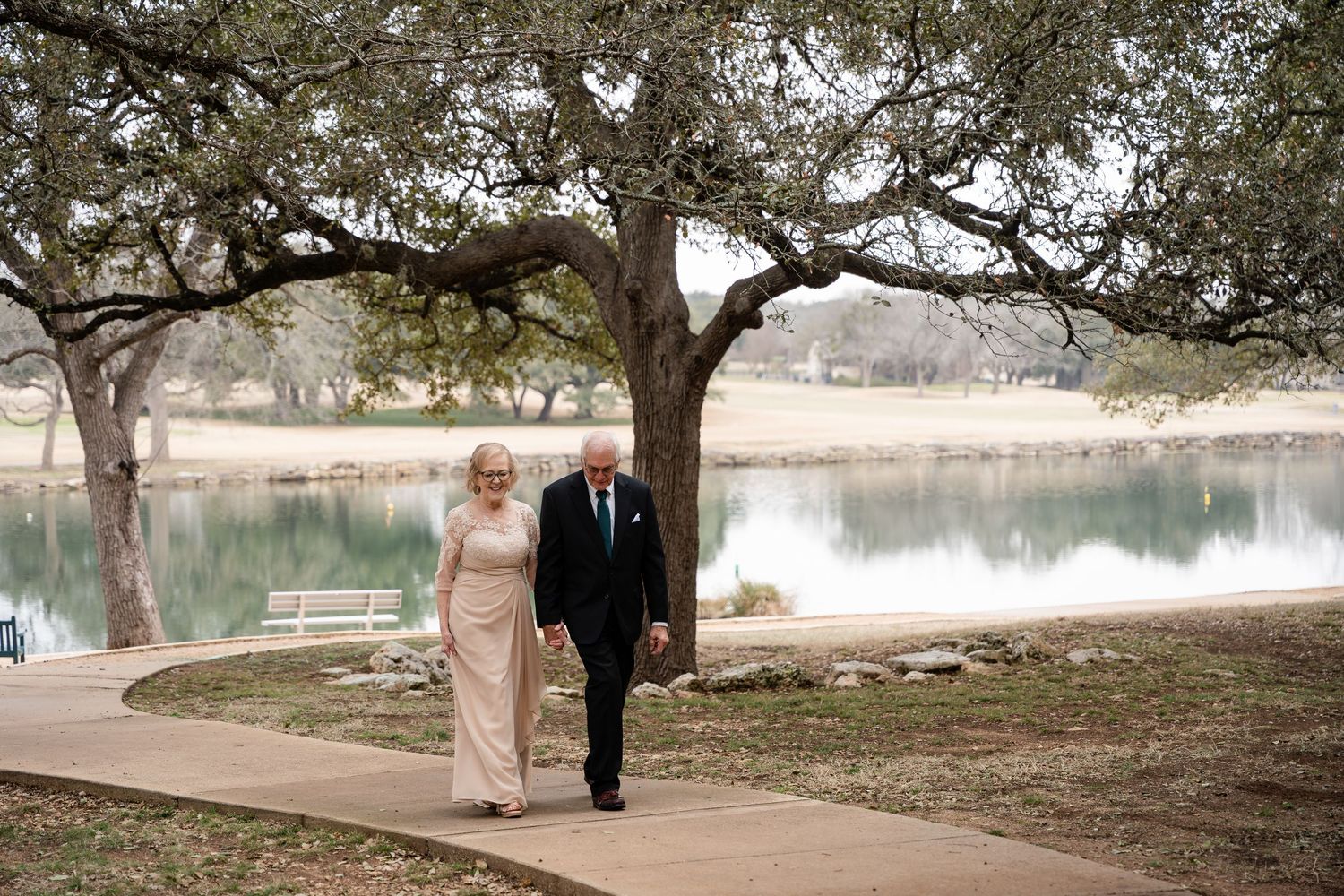Elegant couple walks along scenic lakeside path under sprawling oak trees at outdoor venue.