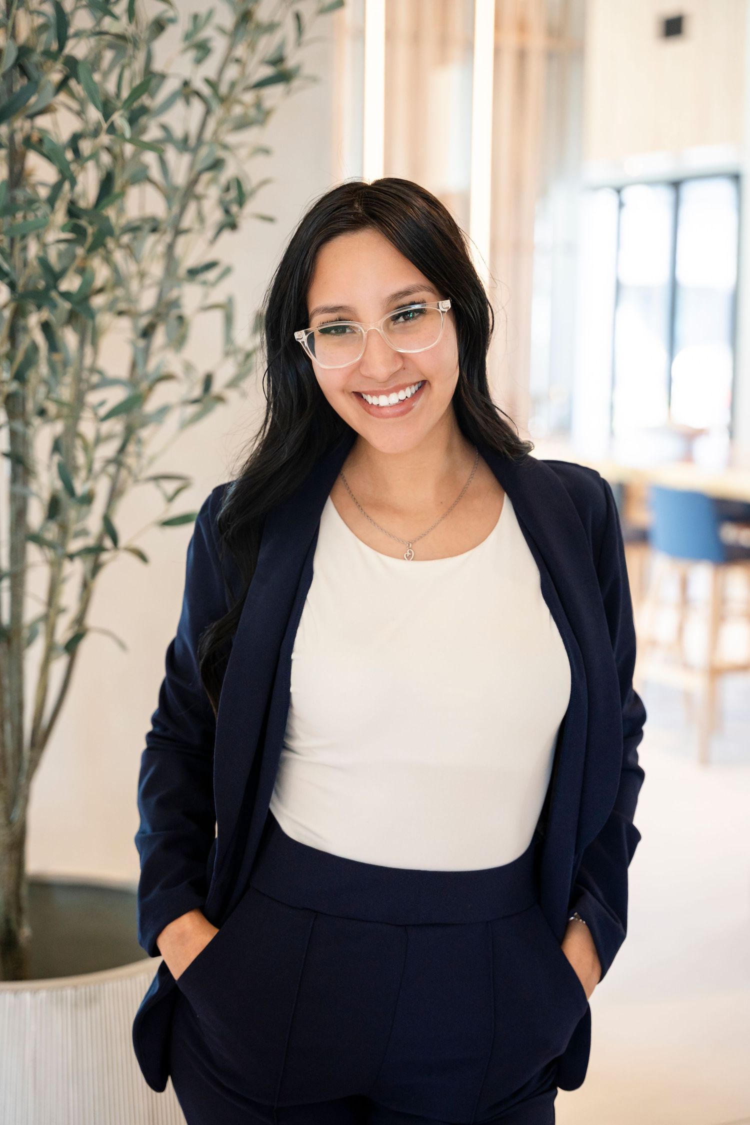 Professional wearing navy blazer and white top stands in bright modern office space with indoor plants.