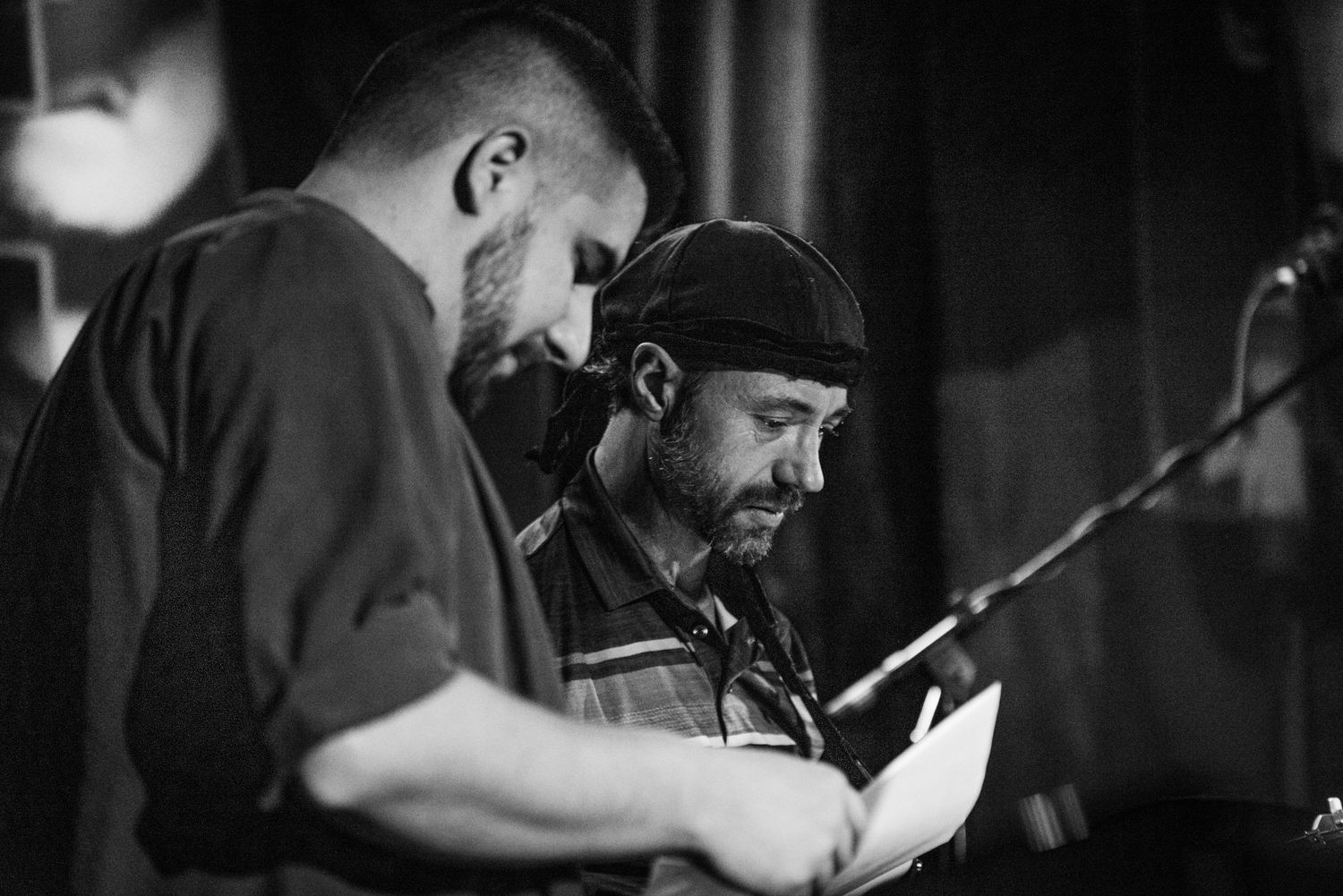Two performers share a moment reviewing sheet music in a monochrome performance setting.