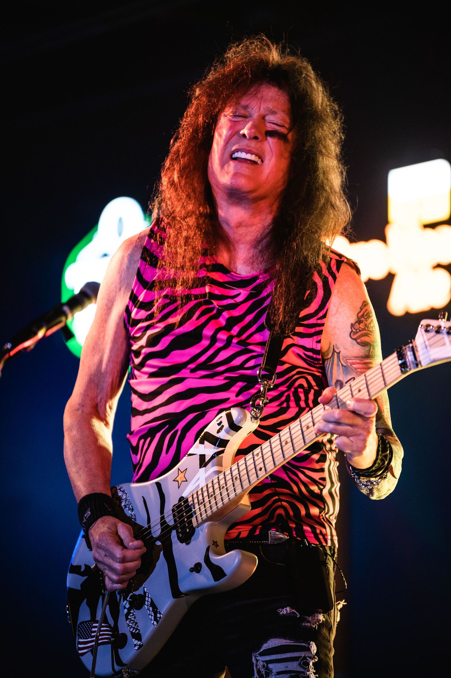 Guitarist plays electric guitar in zebra print tank top under colorful stage lighting.