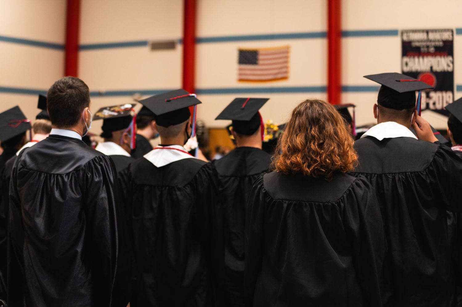 Row of graduates in black caps and gowns standing before American flag indoors.