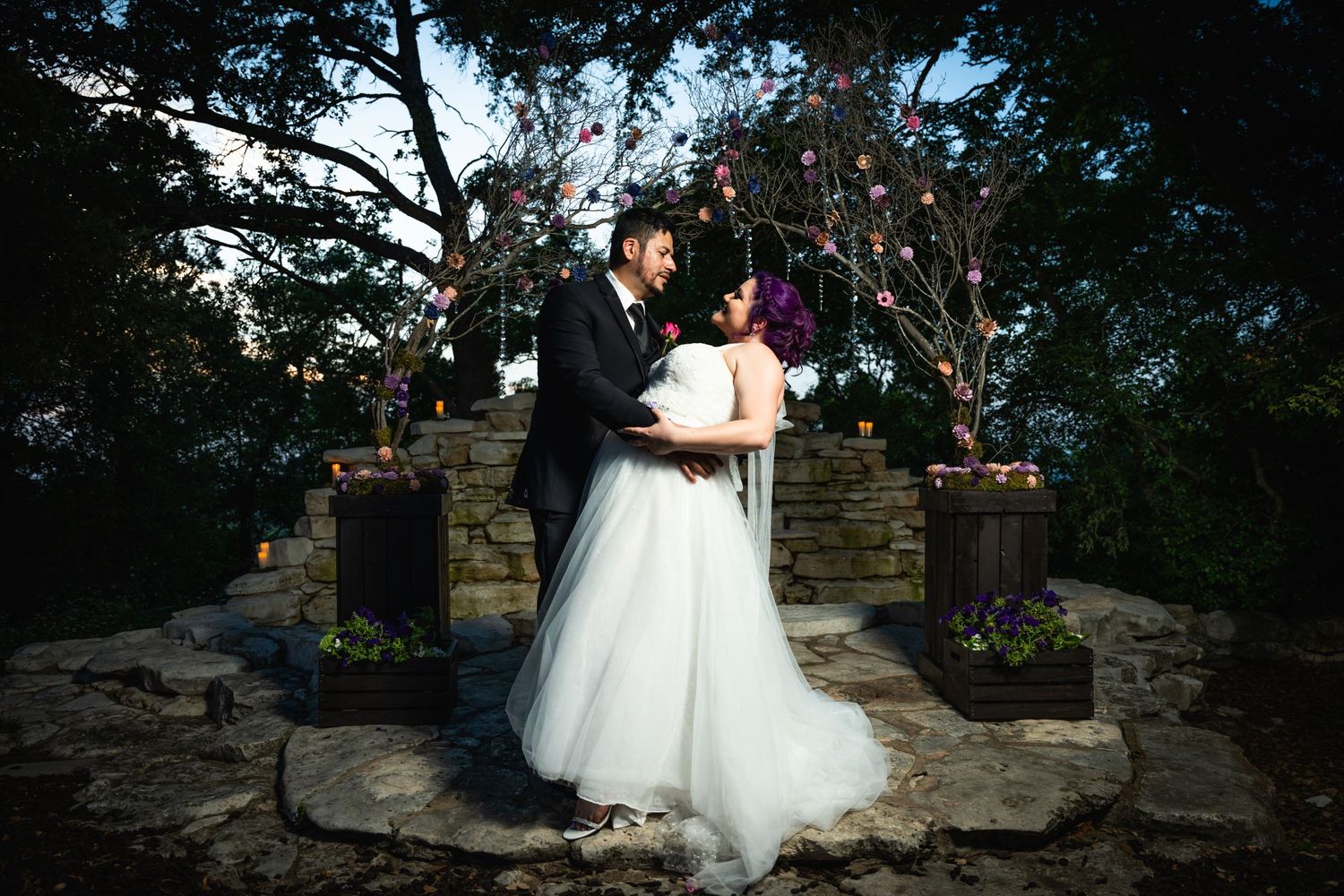 A romantic wedding couple shares an intimate moment at dusk in front of a stone wall decorated with string lights.