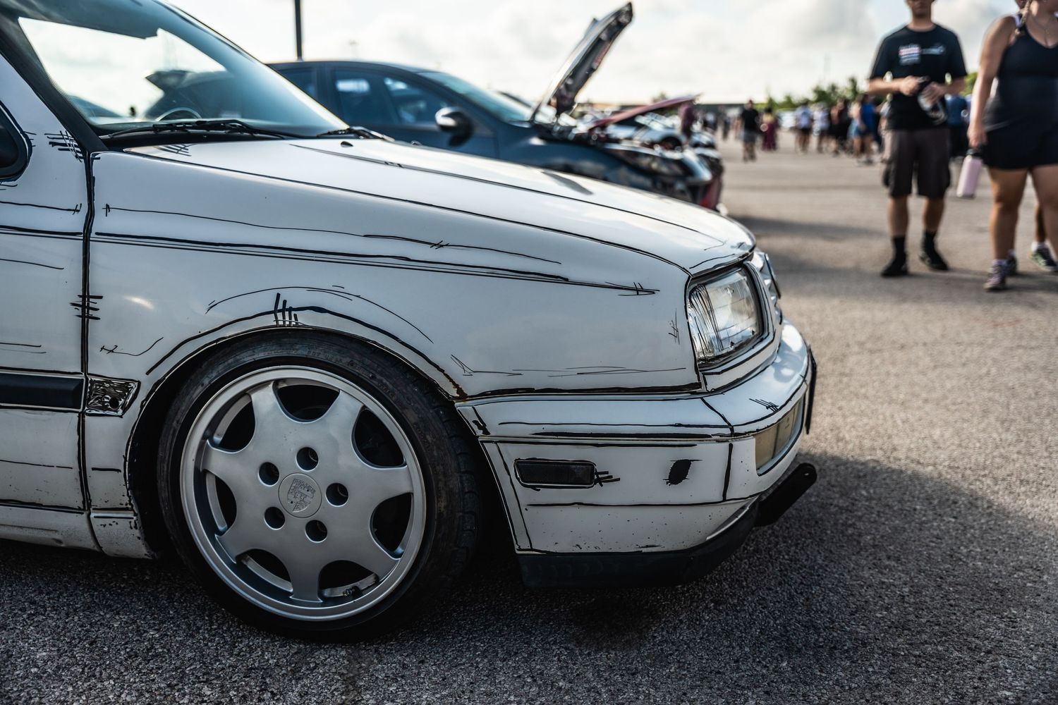 Side profile of a lowered white Volkswagen Golf with custom wheels parked at a car show.