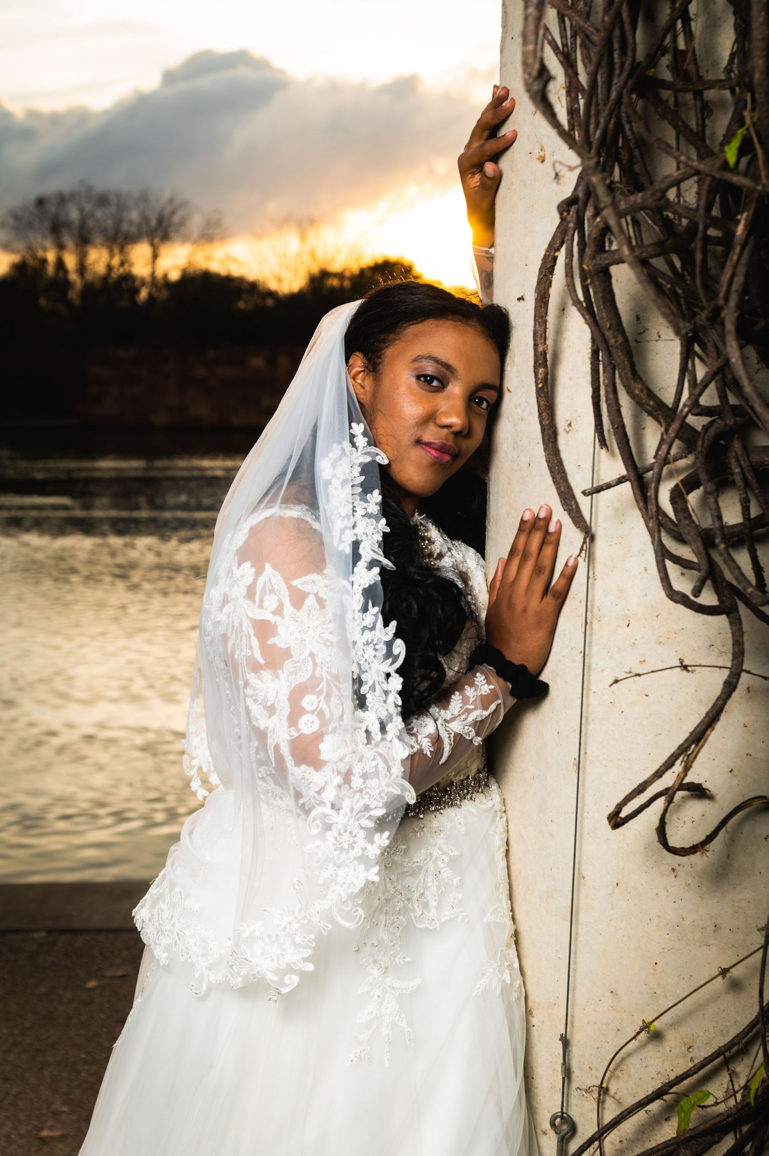 Bride in lace sleeve wedding dress poses against wall at sunset by water.