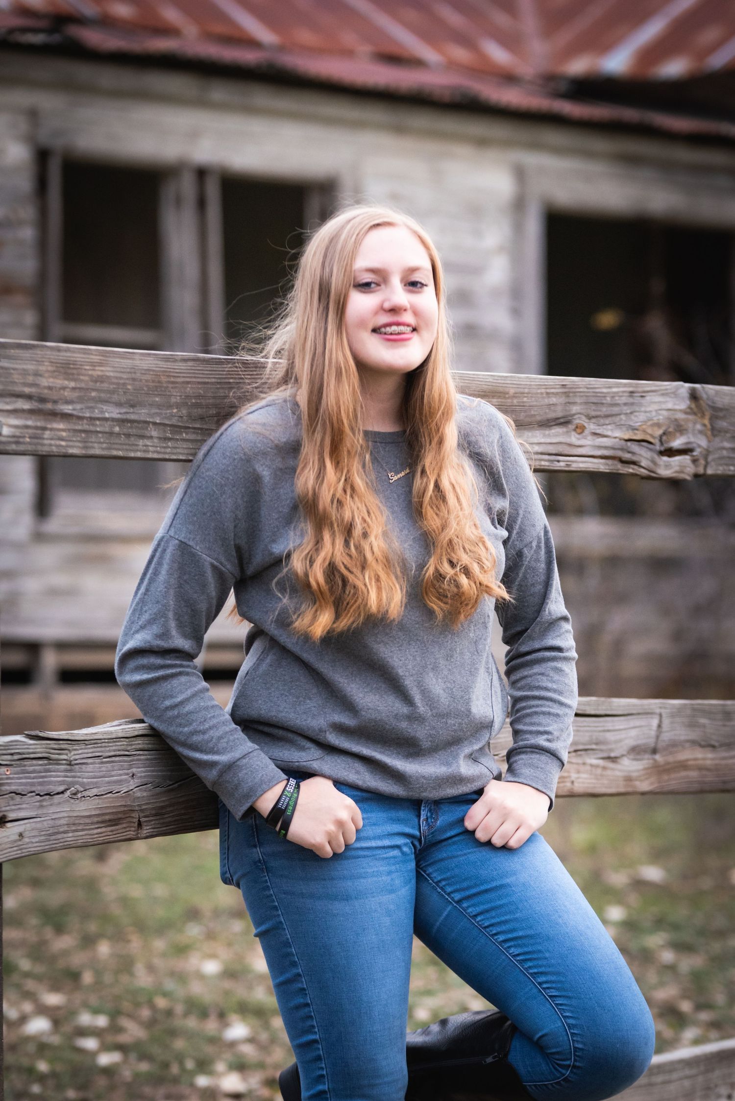 Person in gray sweater and jeans poses casually against rustic wooden building background.