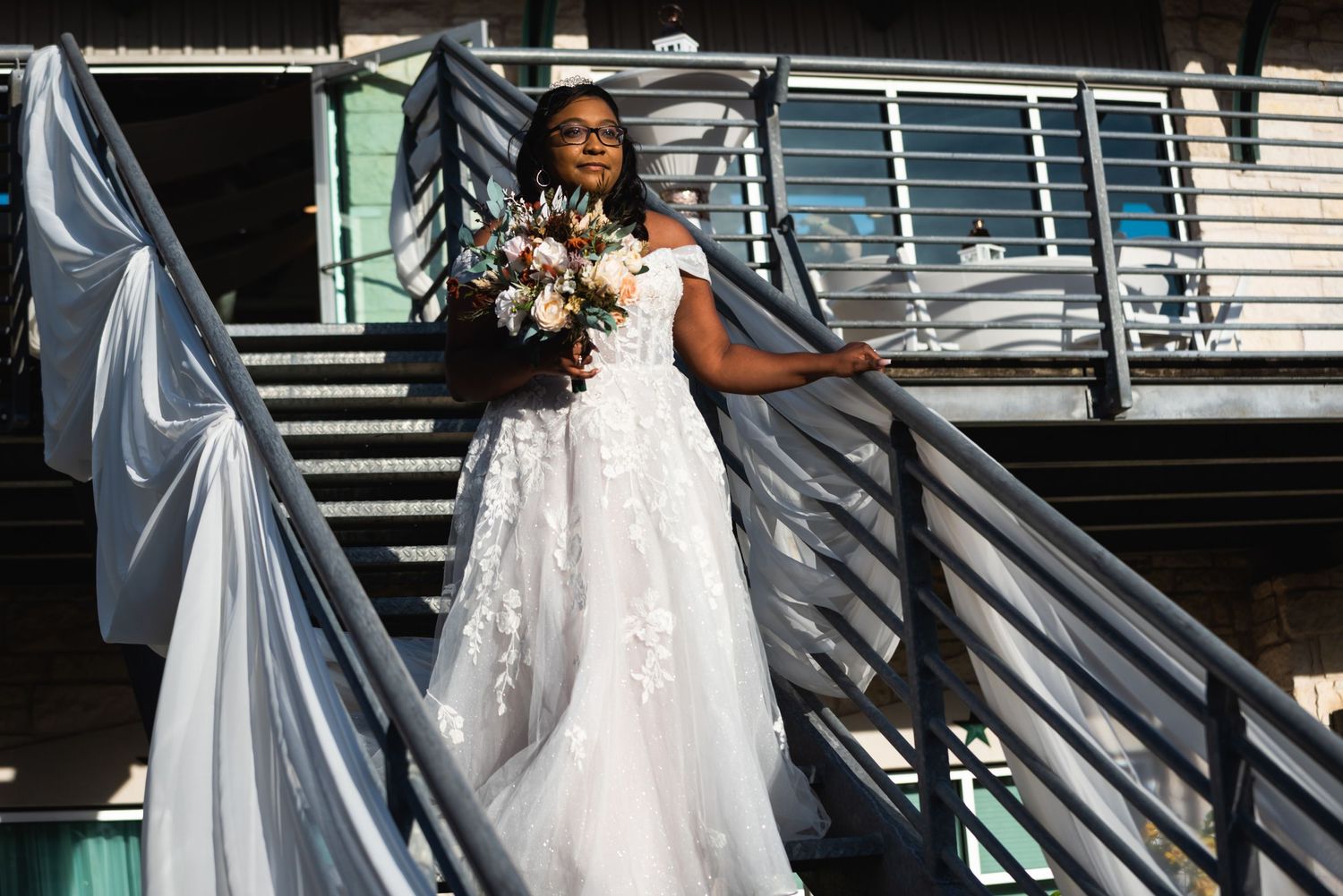 Bride in white lace gown poses on outdoor staircase with flowing bouquet.