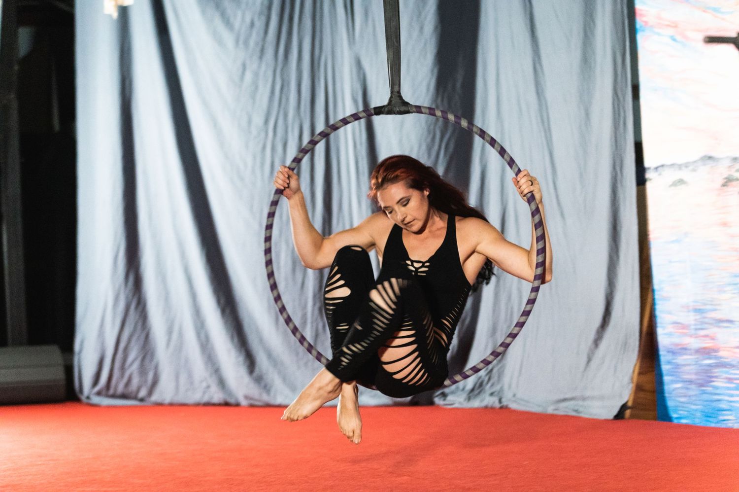 Performer demonstrates control and balance while seated in aerial hoop against grey backdrop.