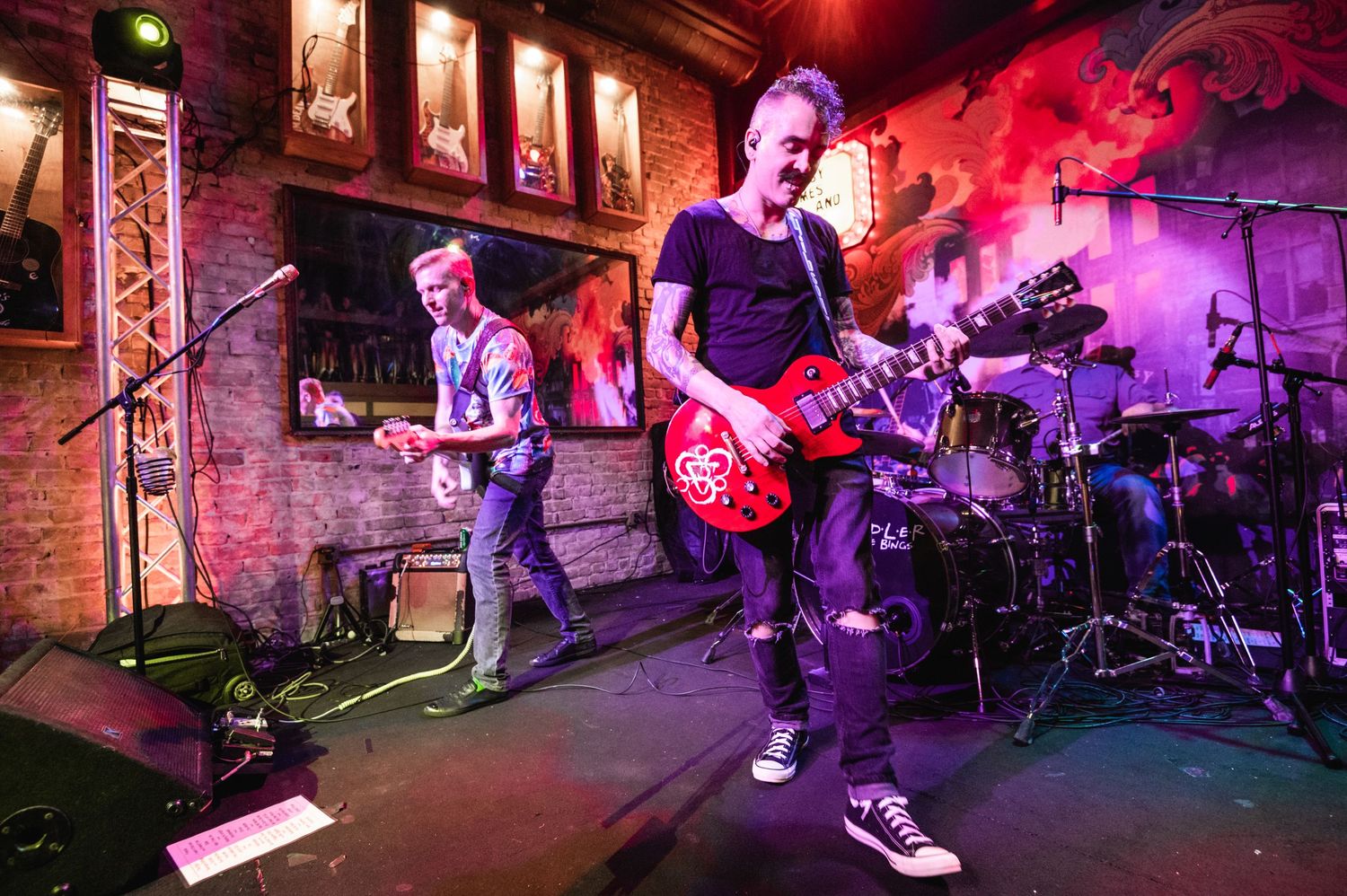Musicians rocking out on stage with red lighting and brick wall backdrop.