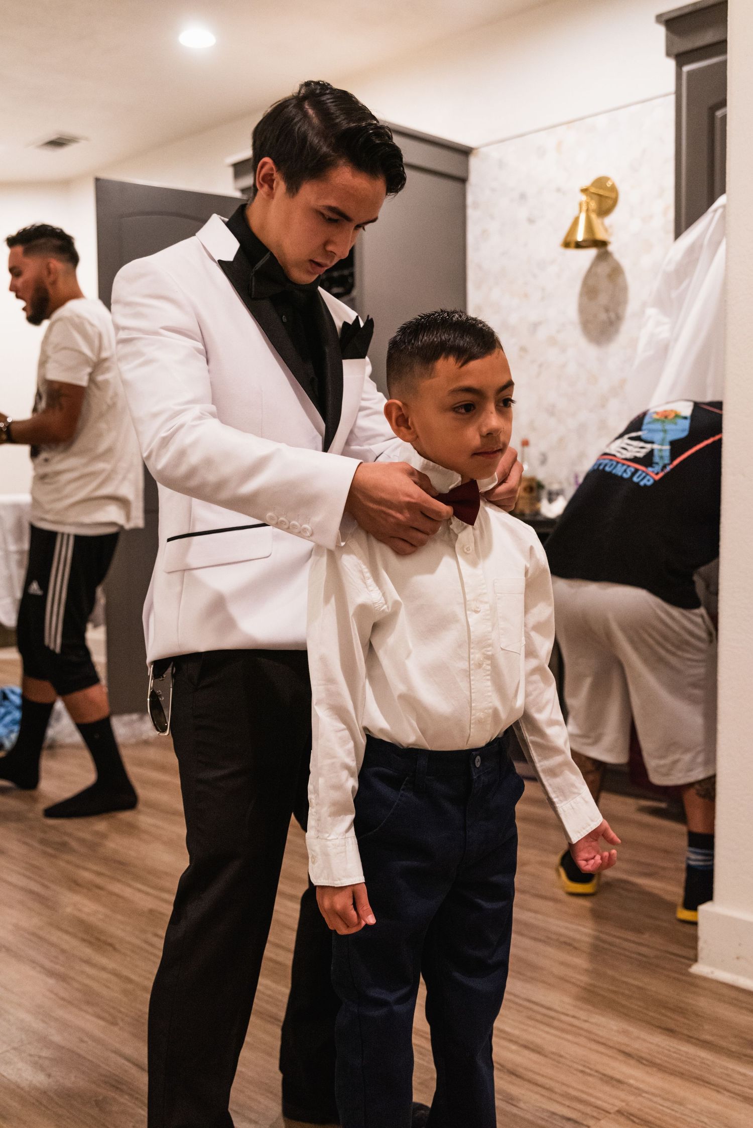 A formal bowtie adjustment during wedding preparations with a white tuxedo jacket and dark dress pants.