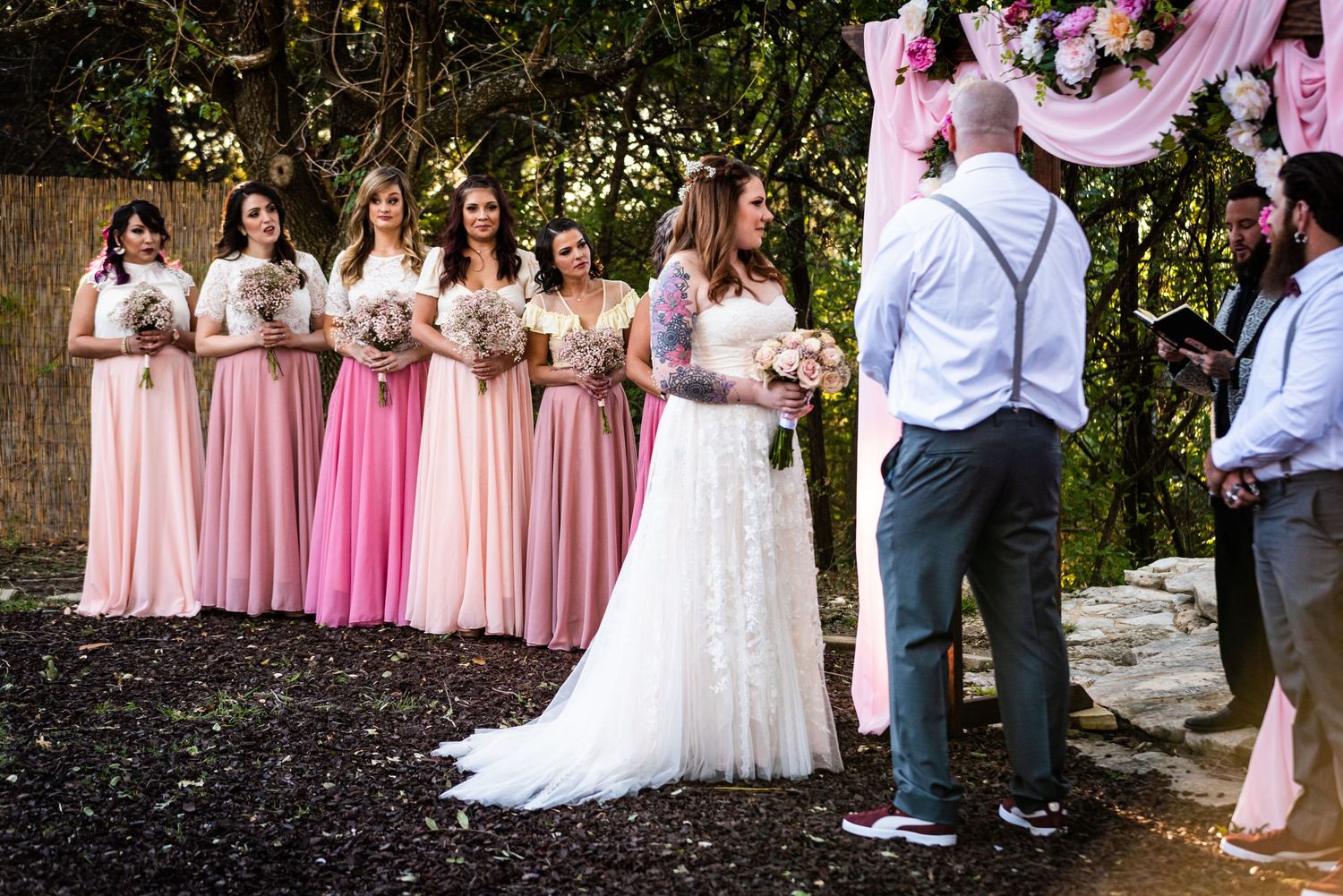Bridesmaids in pink ombre dresses line up during outdoor wedding ceremony with floral arch.