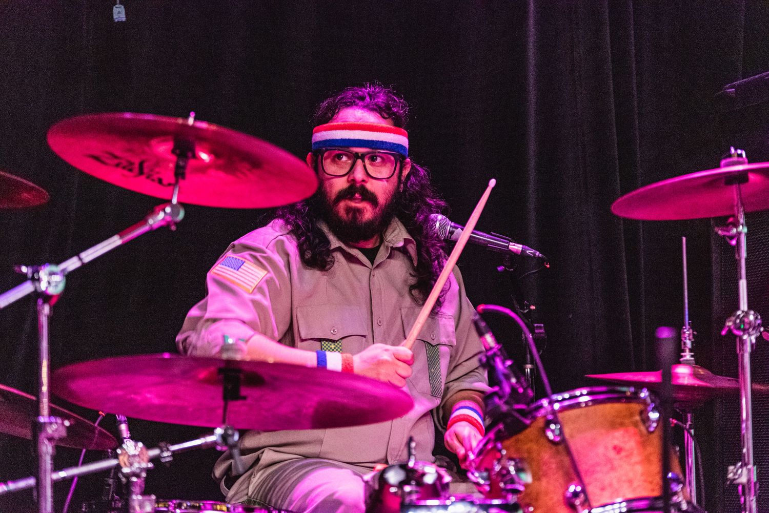 Focused drummer wearing glasses and uniform performs behind drum kit under magenta lighting.