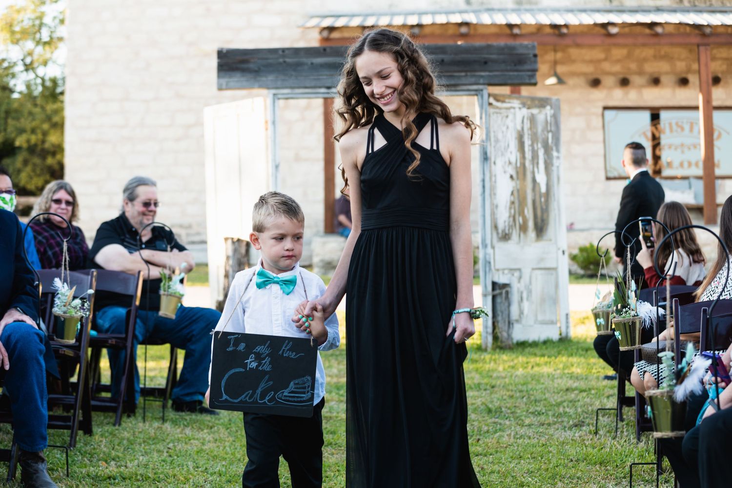 Wedding party member in black dress walks with young ring bearer in bow tie during ceremony.