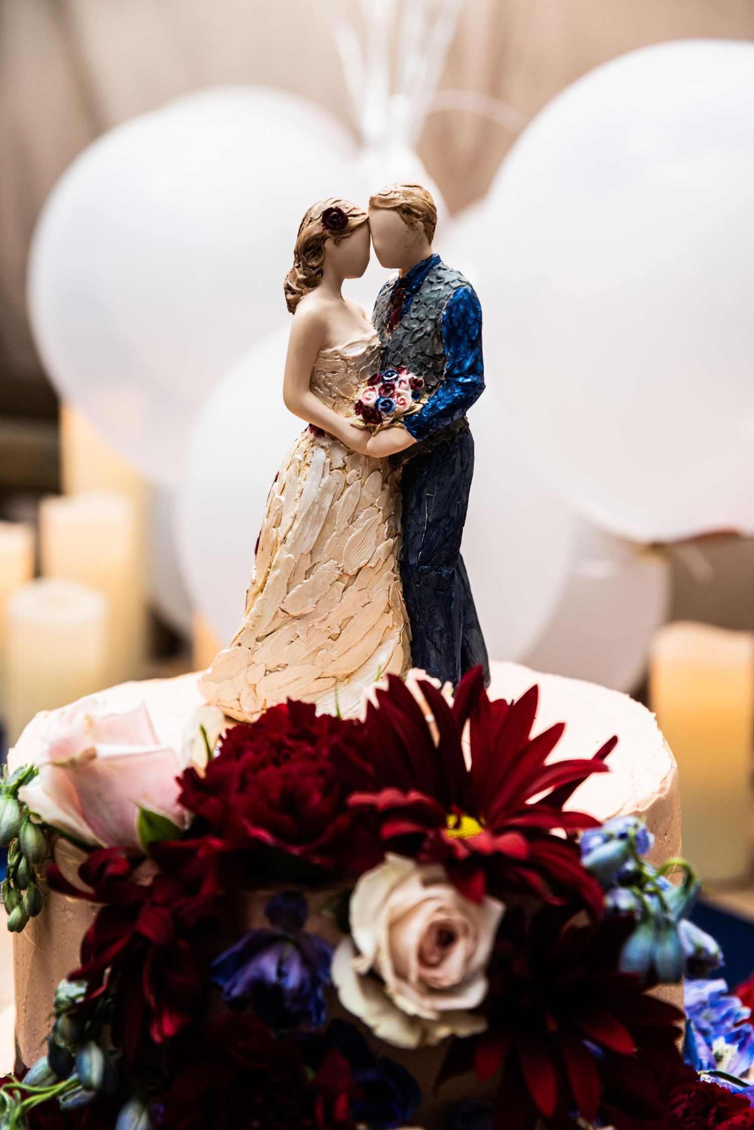 Close-up of wedding cake topper figurines surrounded by burgundy flowers and white balloons.