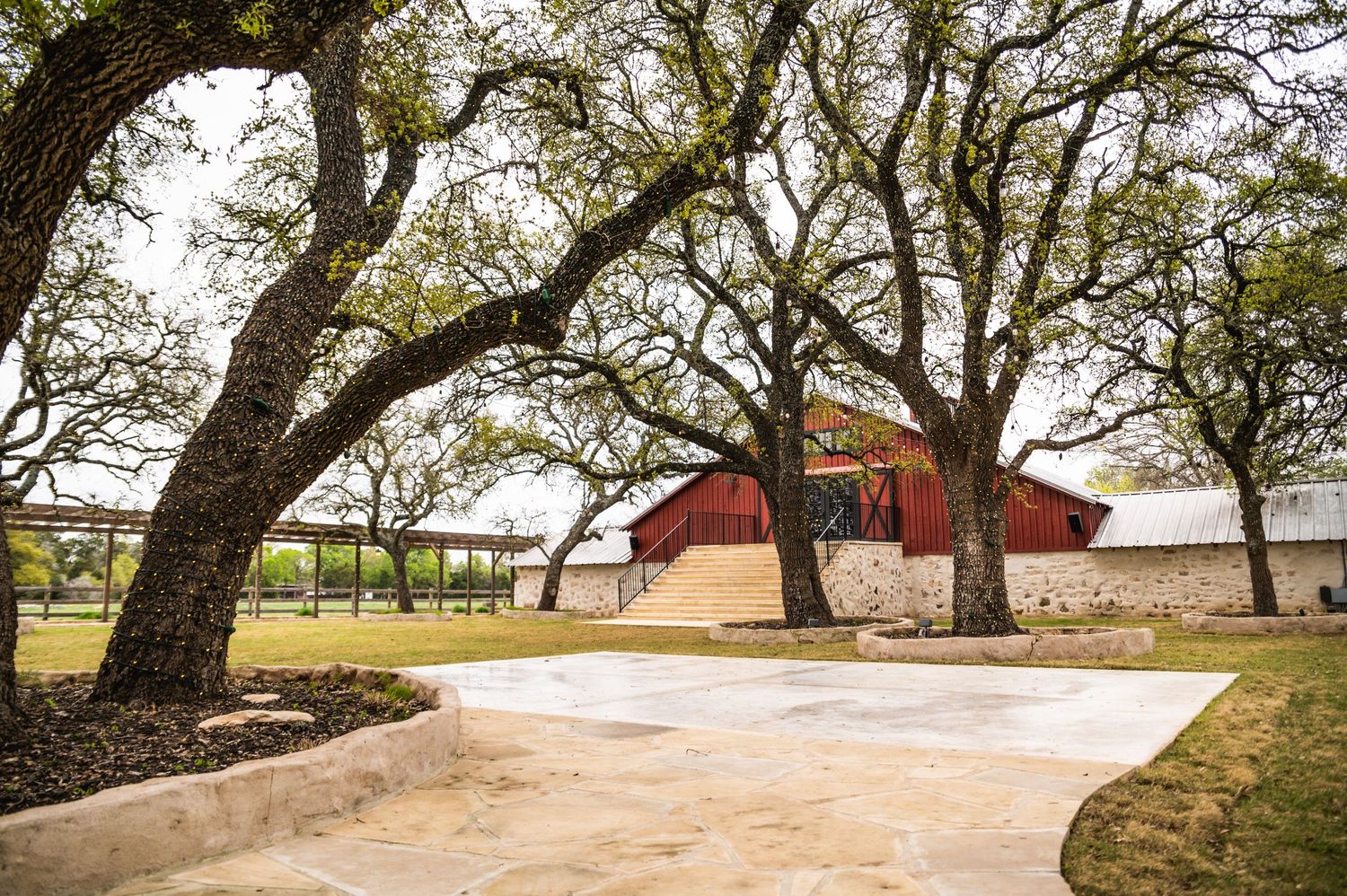 A rustic red barn building with large oak trees framing the driveway entrance.