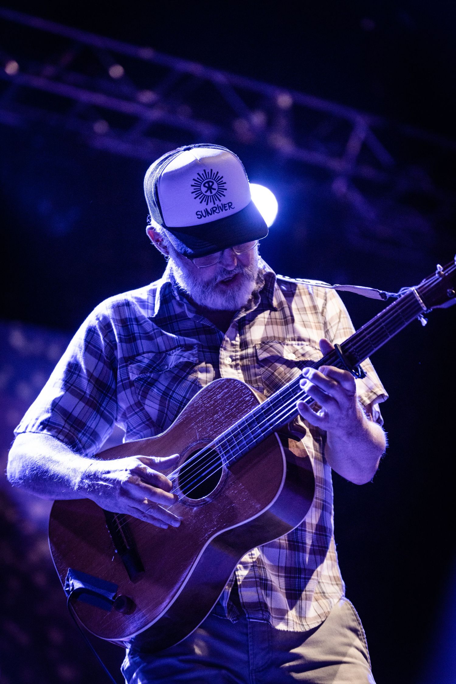 Musician in baseball cap playing acoustic guitar under purple stage lighting.