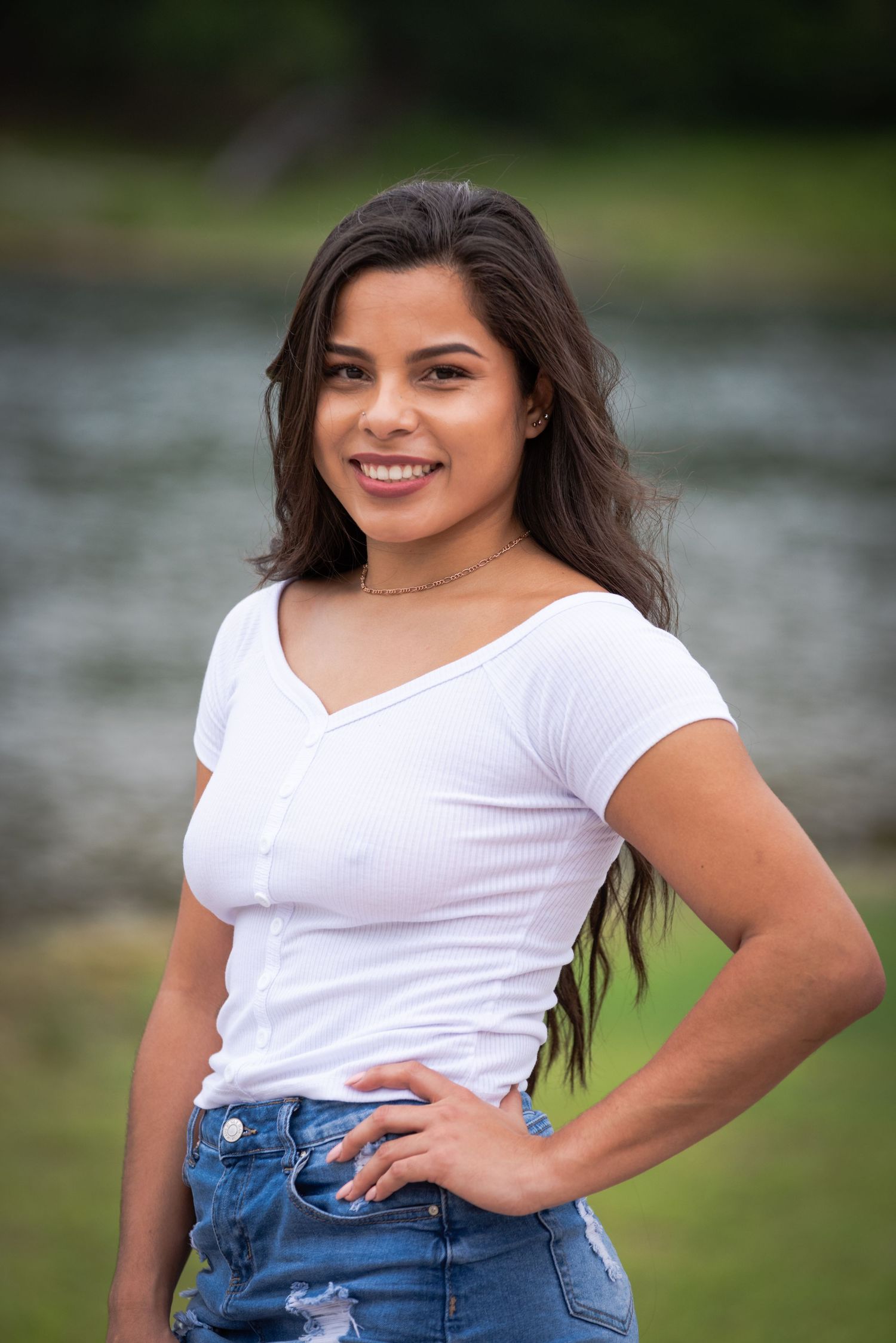 A casual portrait of a young woman in a white top and ripped jeans standing by a lake.