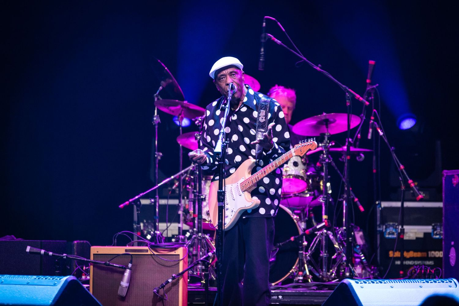 Musician in polka dot shirt and white cap performs on stage with dramatic purple and blue lighting.