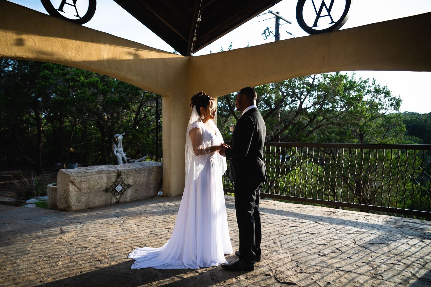 Couple in wedding attire stands together on stone patio under covered pavilion.