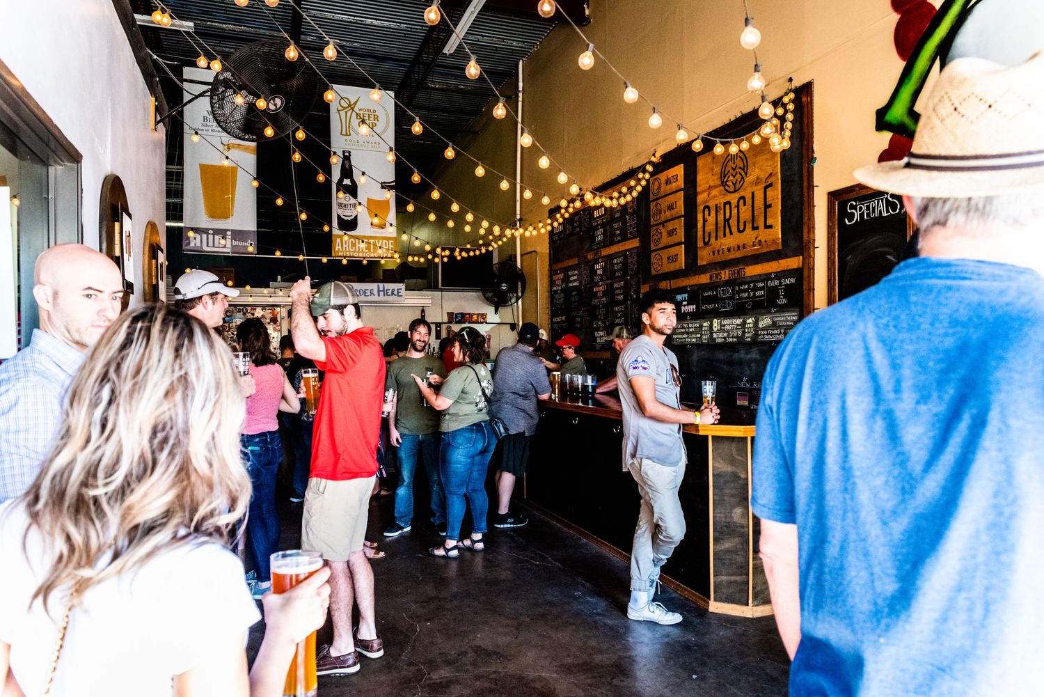 Interior of a craft brewery taproom with string lights and customers enjoying beverages.
