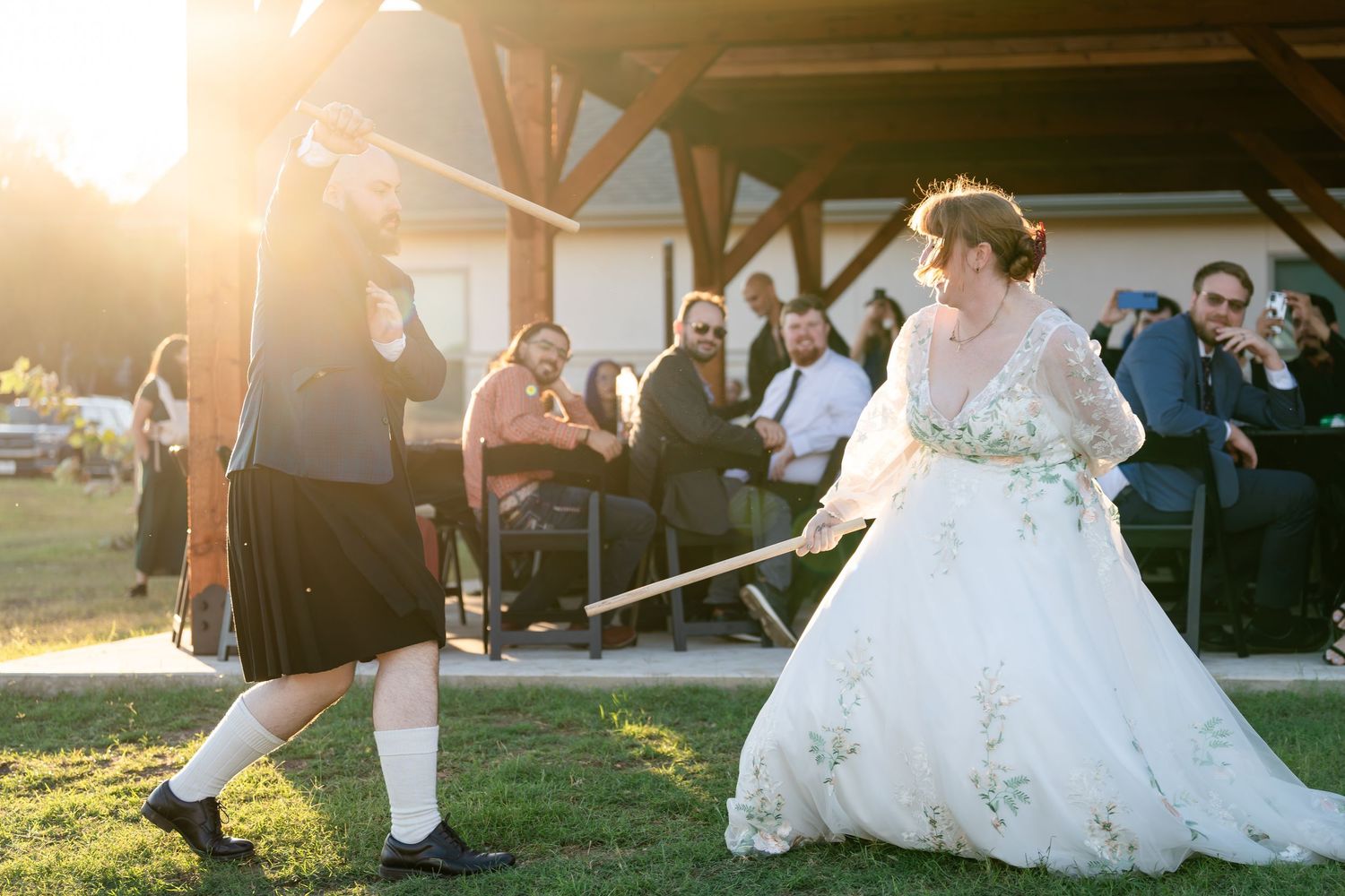 A bride in a white lace wedding dress dances joyfully at an outdoor sunset reception with guests watching.