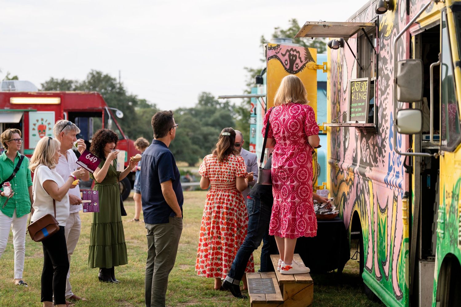Customers in summer dresses wait in line at a colorful food truck at an outdoor festival.
