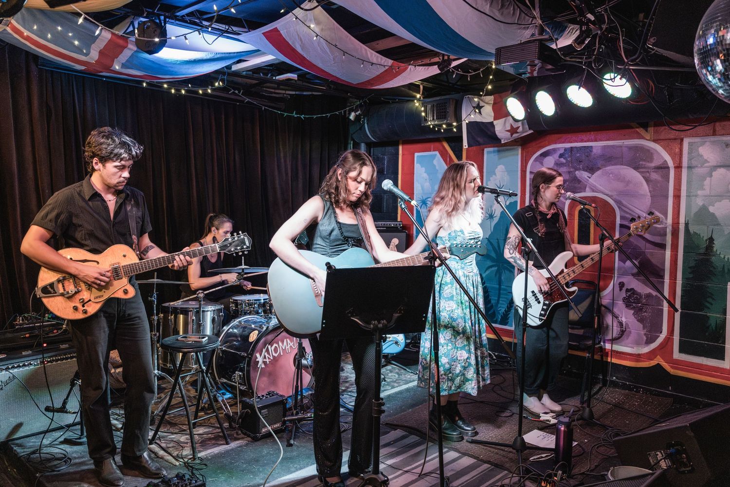 Musicians perform on stage at a live music venue with instruments and stage lighting visible in the background.