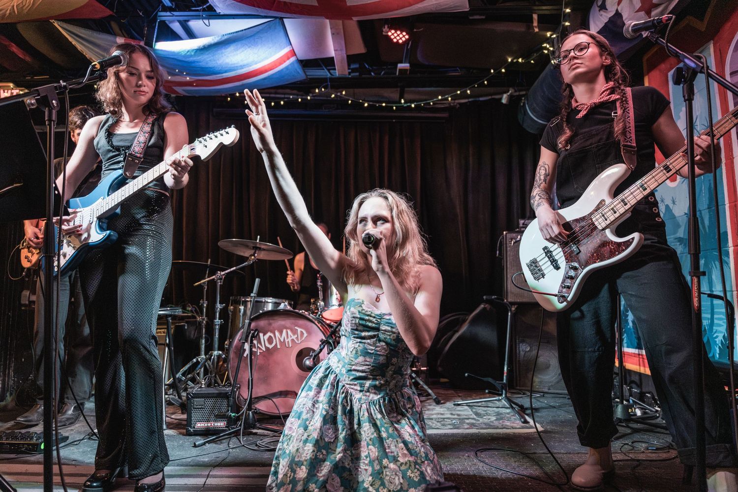 A rock band performs on stage with two guitarists and a singer in a blue dress under moody lighting at a live venue.