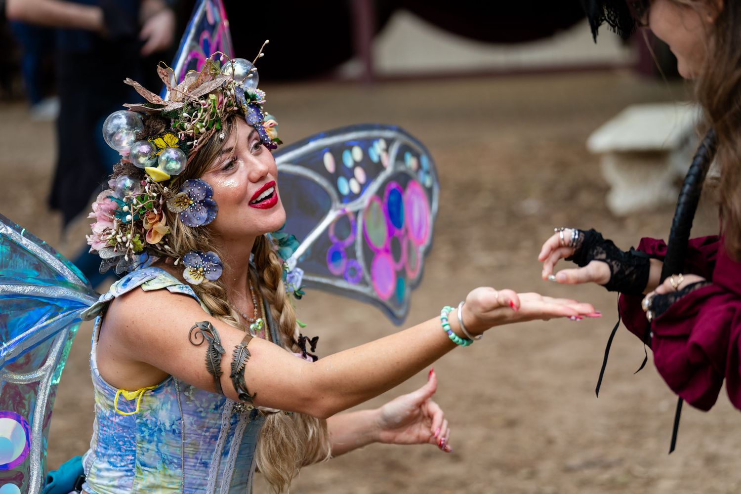 A performer in colorful fairy wings and festival attire dances with bubbles at an outdoor event.