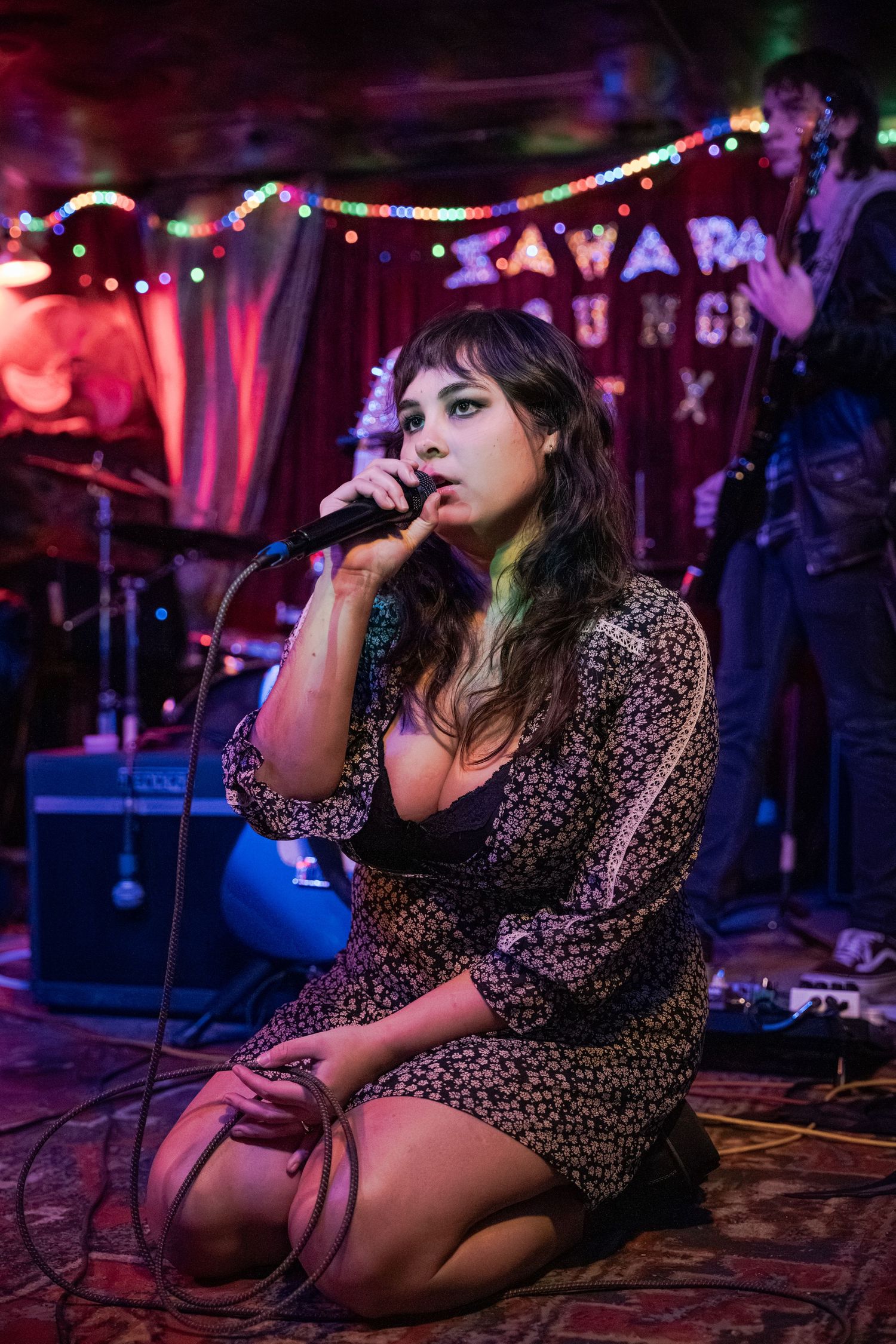 Singer in a floral dress performs on stage under moody purple lighting at a live music venue.