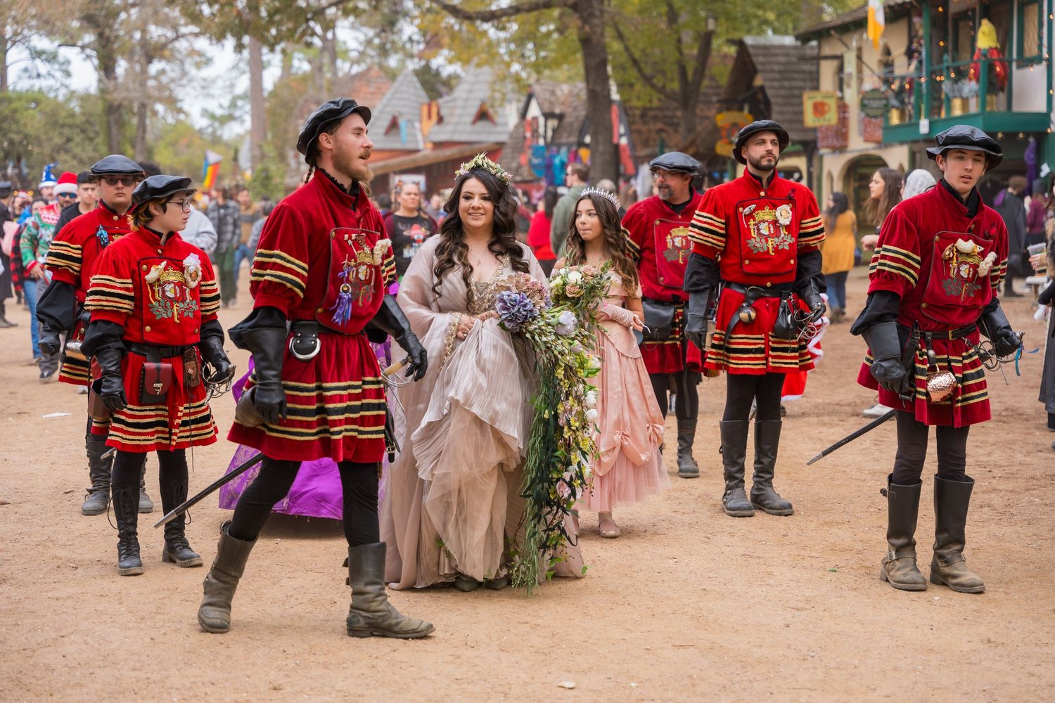 Medieval costumed performers in red and black outfits escort elegantly dressed attendees at a Renaissance festival.