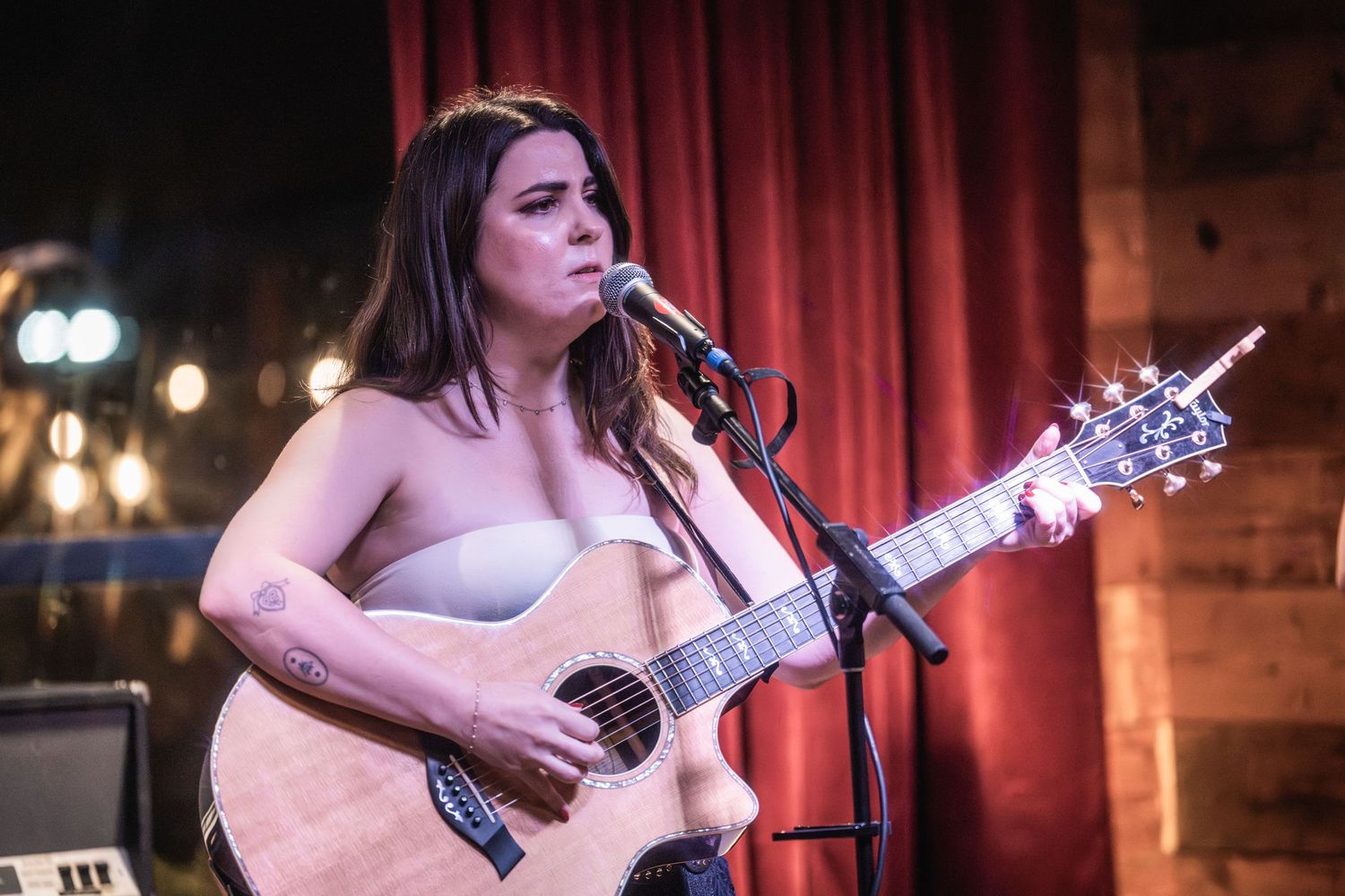 A musician performs on stage with an acoustic guitar in front of a red curtain backdrop under warm lighting.
