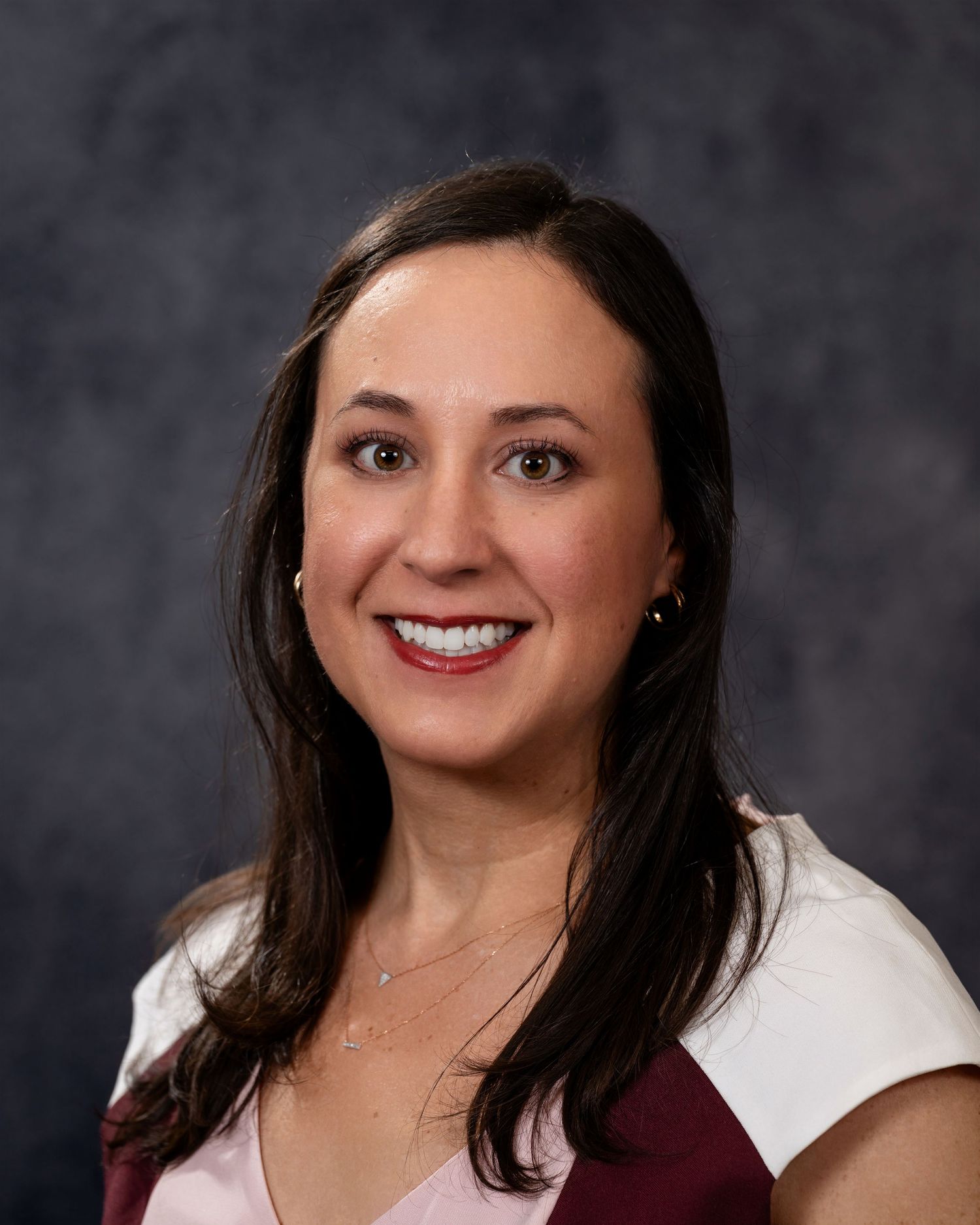 Professional headshot portrait of someone wearing a white and burgundy top against a dark gray background.