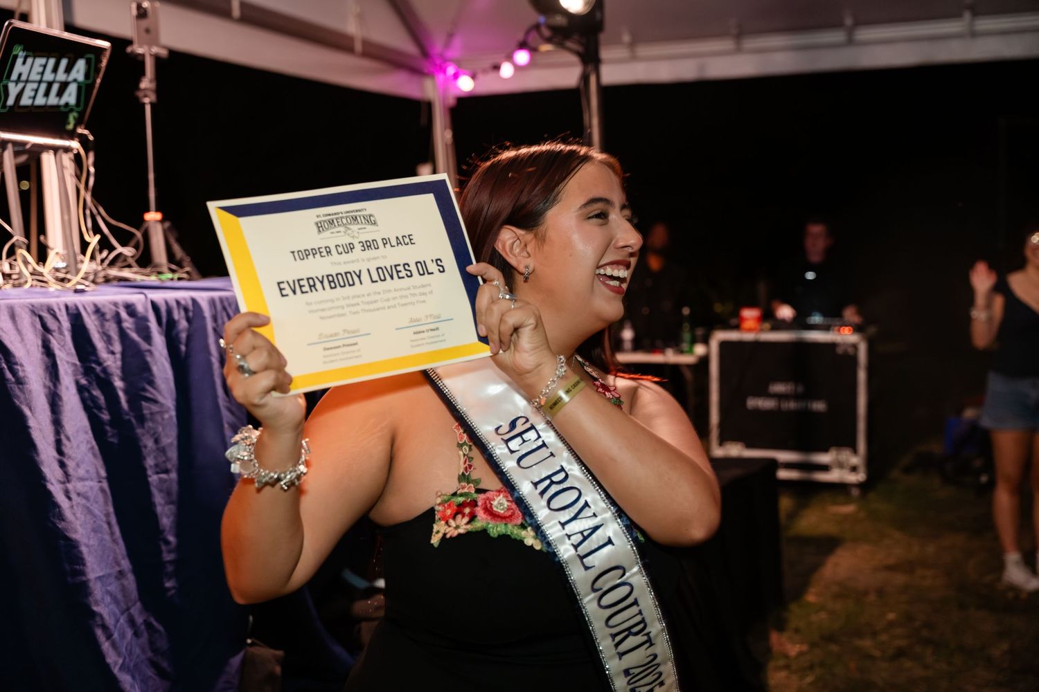 A group of pageant contestants laugh and celebrate together during a nighttime event with purple lighting.