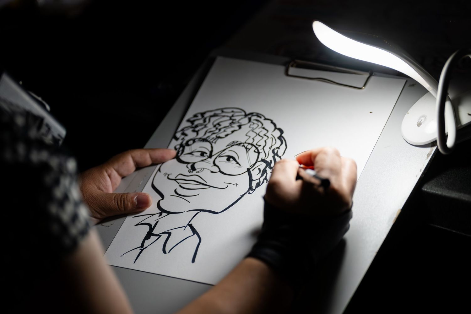 Artist sketches a portrait illustration on white paper under desk lamp lighting in dramatic black and white.