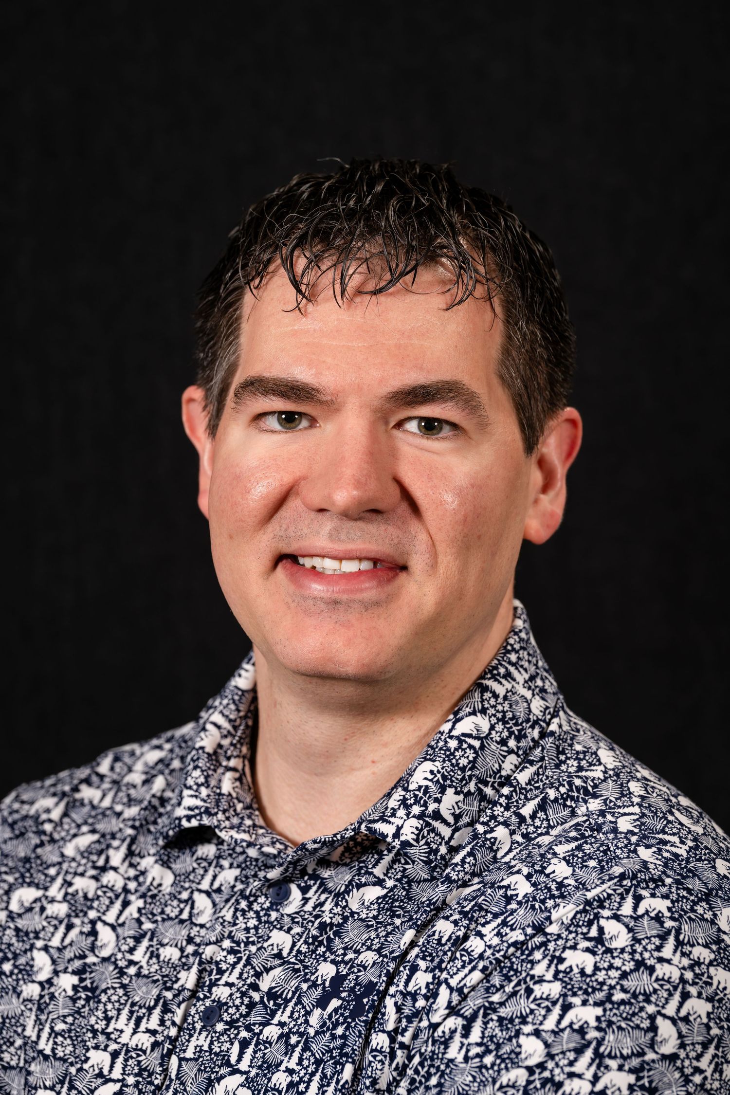 A person wearing a black and white floral patterned shirt poses against a dark background in a professional headshot.
