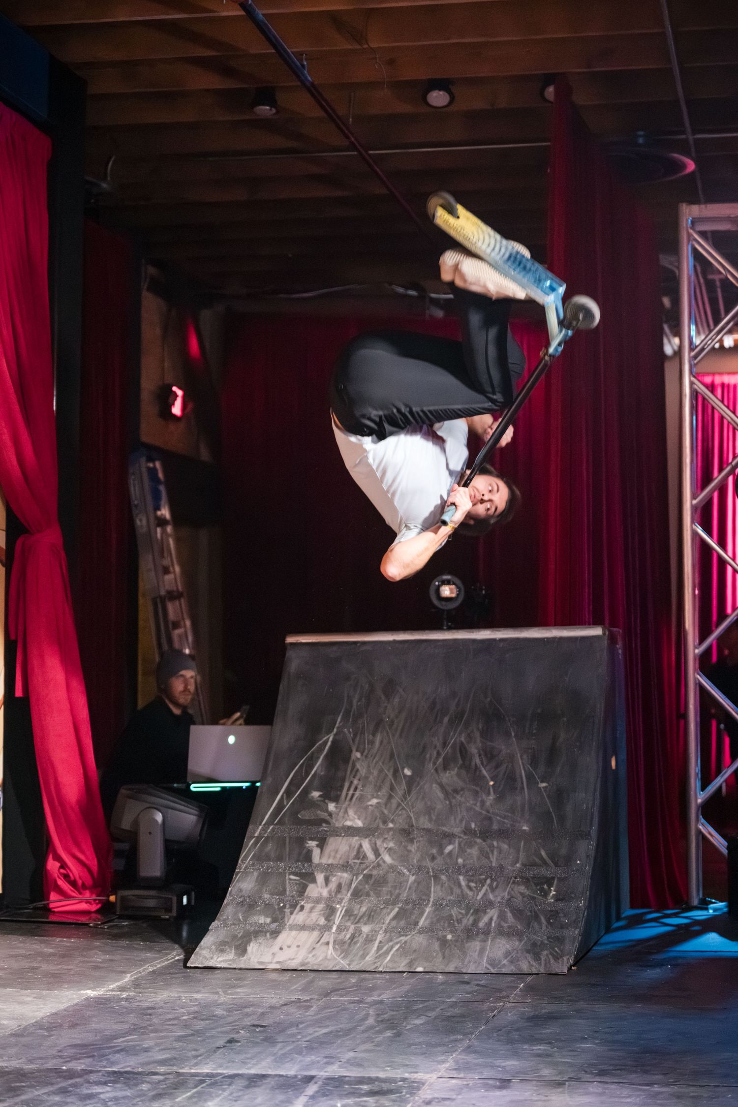 Skateboarder performs an aerial trick sequence on a curved ramp in an indoor skate park with red curtains.