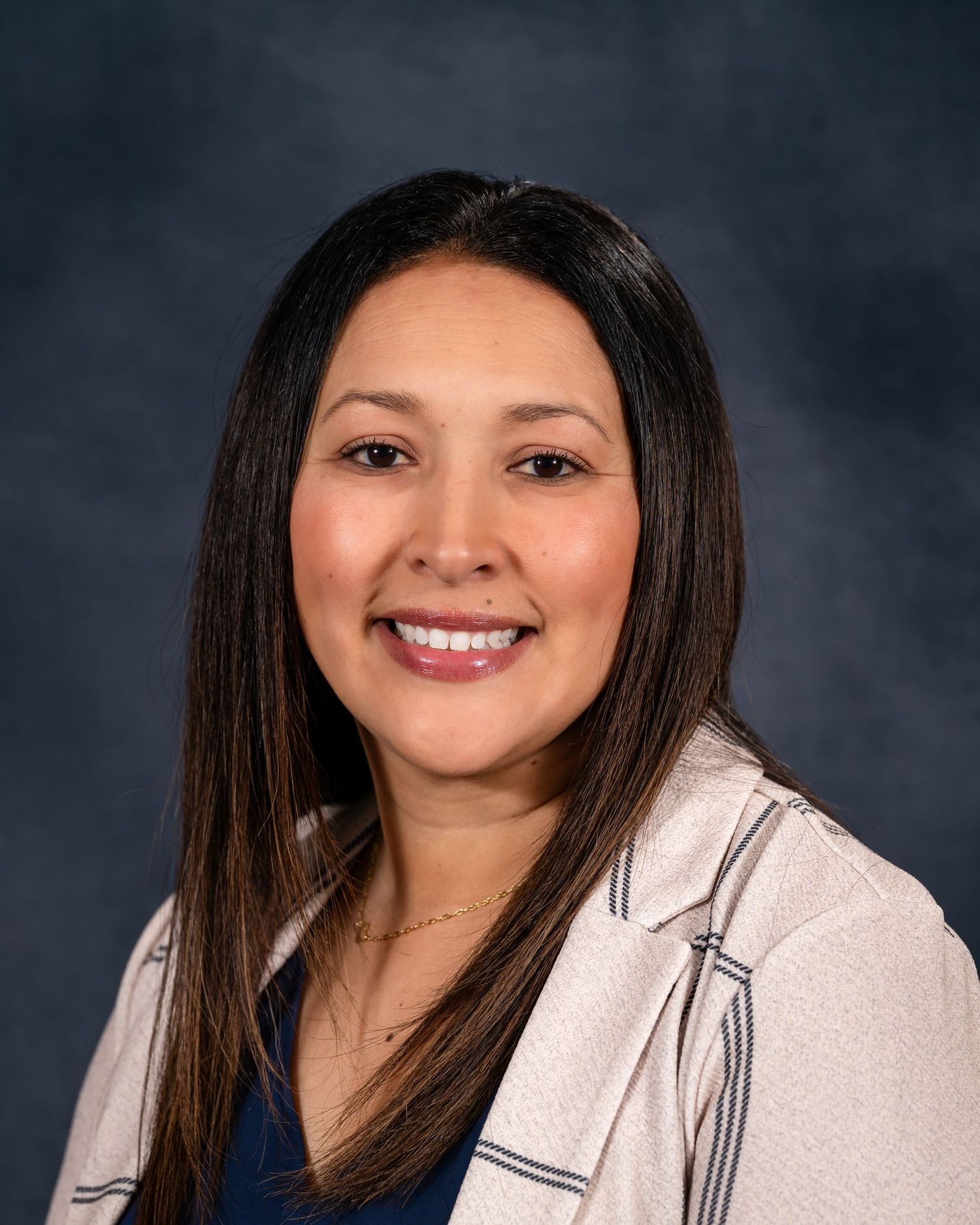 Professional headshot of a person in a beige blazer and dark top against a mottled blue background.