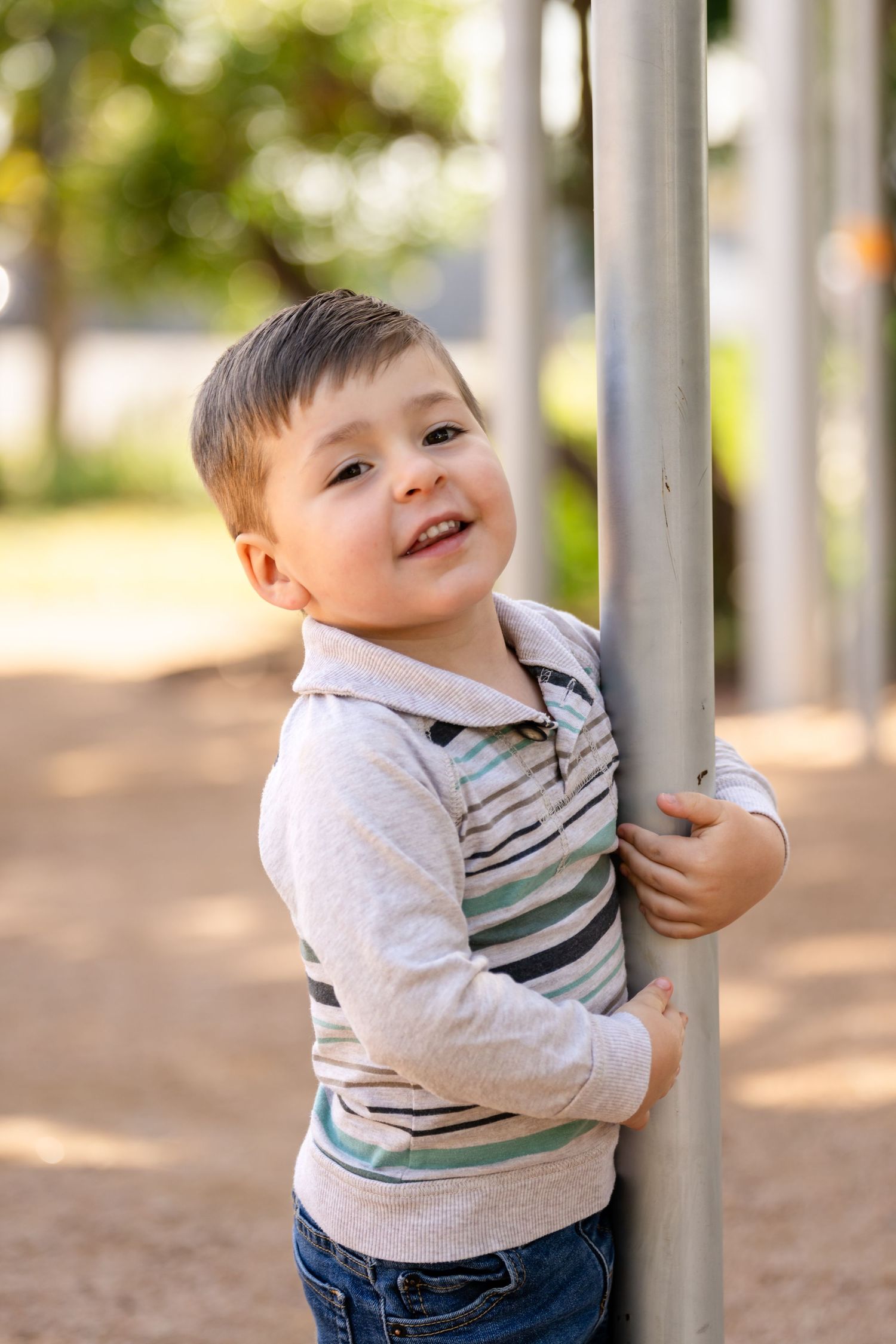 A young child in a striped shirt smiles and plays on metal playground equipment on a sunny day.