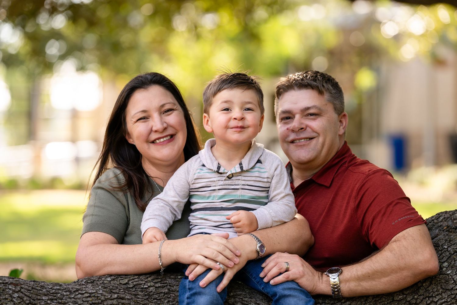 A family poses together outdoors on a sunny day while wearing casual clothing in a park setting.