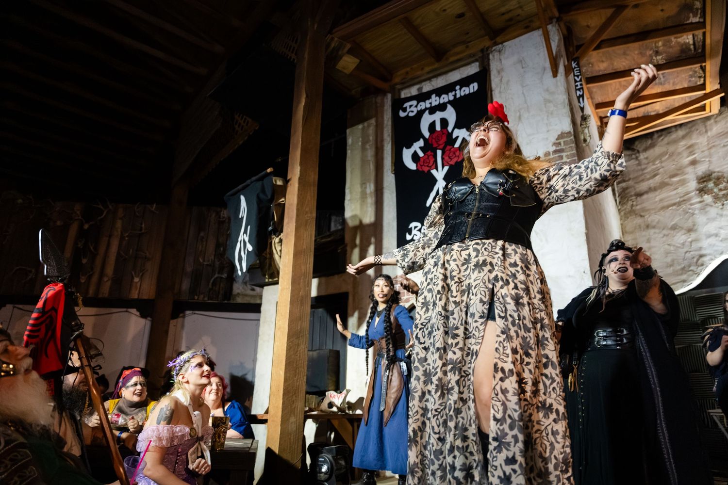 A performer in a floral dress gestures dramatically during a theatrical performance in an industrial warehouse space.