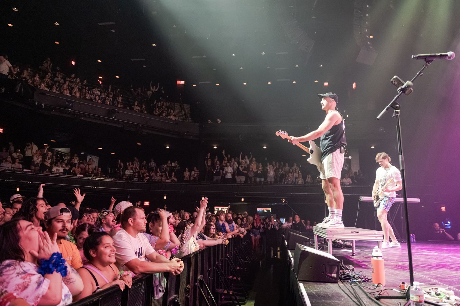 A rock band performs on stage before a packed crowd in a large indoor concert venue with balcony seating.